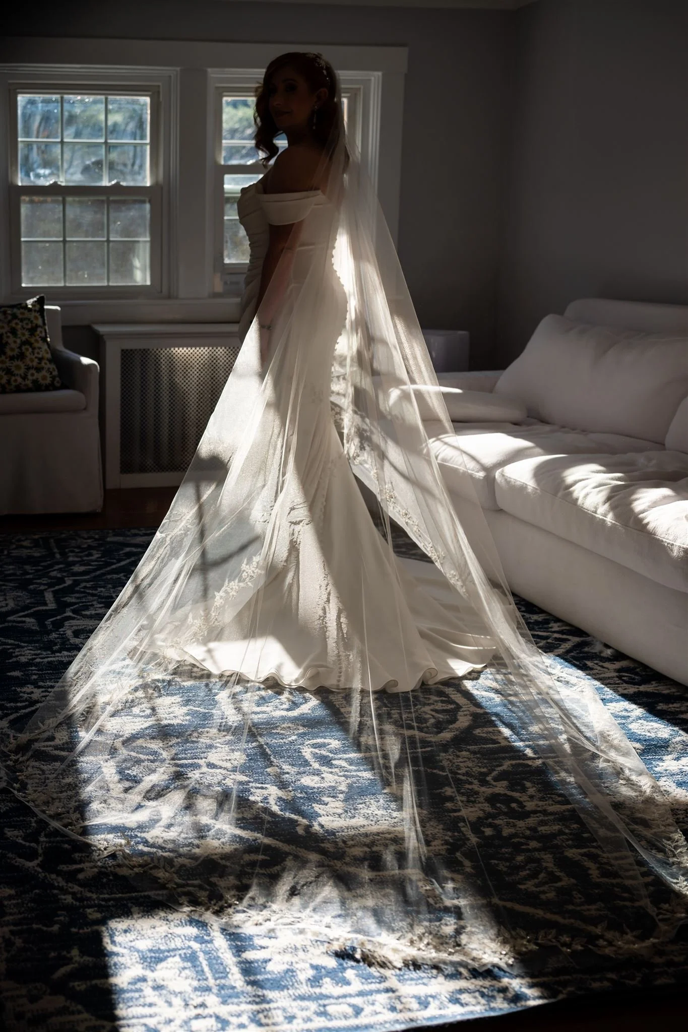 A woman in a white wedding dress with a long, flowing veil standing in a room with large windows, sunlight shining through, with a couch and patterned carpet visible at Lambert's Cove Inn on Martha's Vineyard.