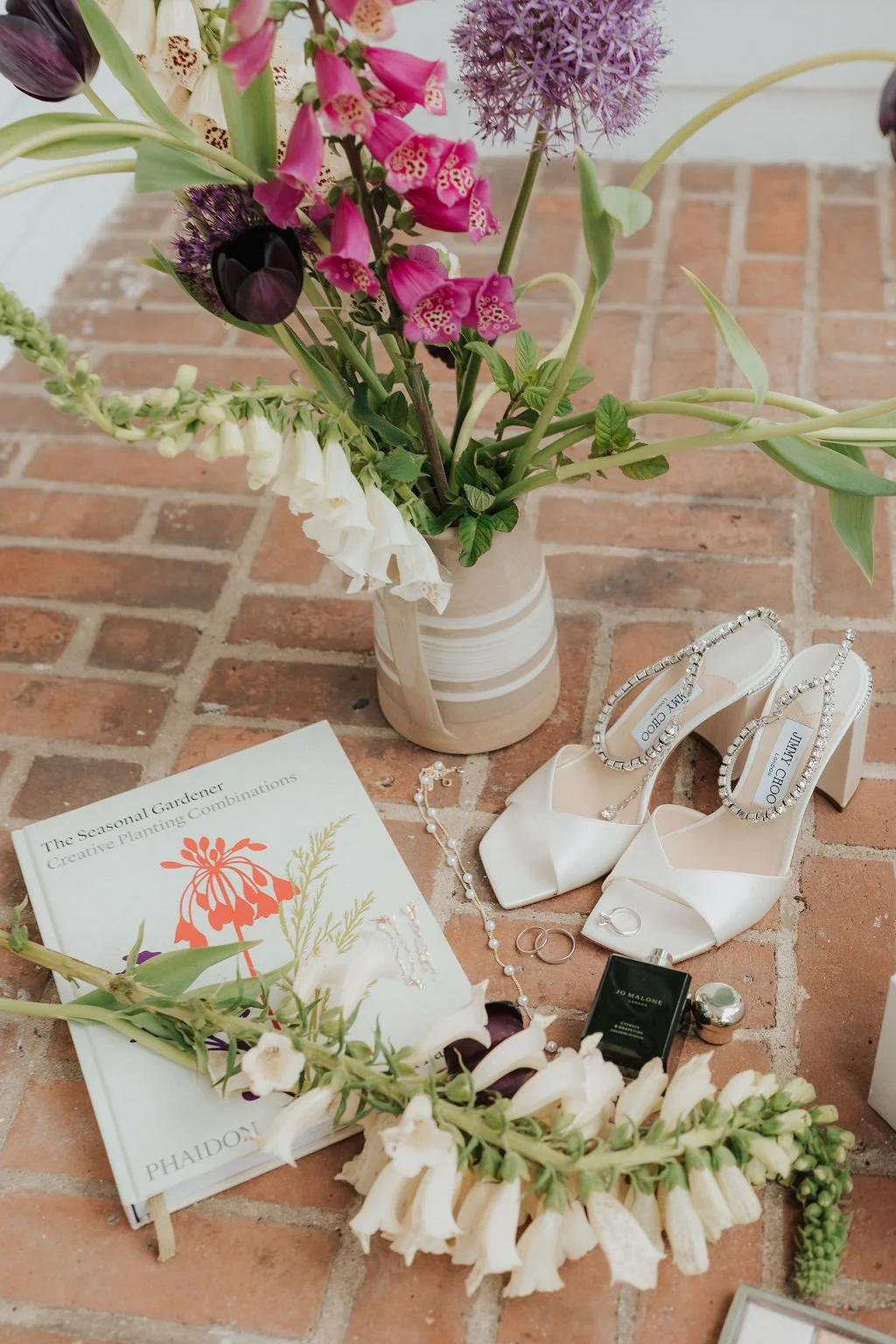 A bouquet of various flowers, a pair of white high-heeled shoes with rhinestone straps, jewelry including earrings, rings, a necklace, perfume, and a book about creative planting arrangements, all arranged on a brick surface.
