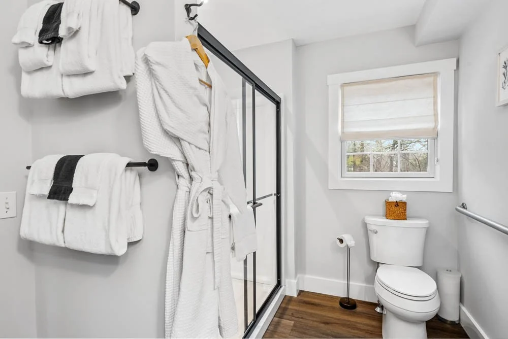 Bathroom with white walls, a window with a blind, a toilet, and a glass shower enclosure. White towels hang on wall-mounted bars, and a white bathrobe is on a hook. Wood flooring and a small basket on top of the toilet tank.