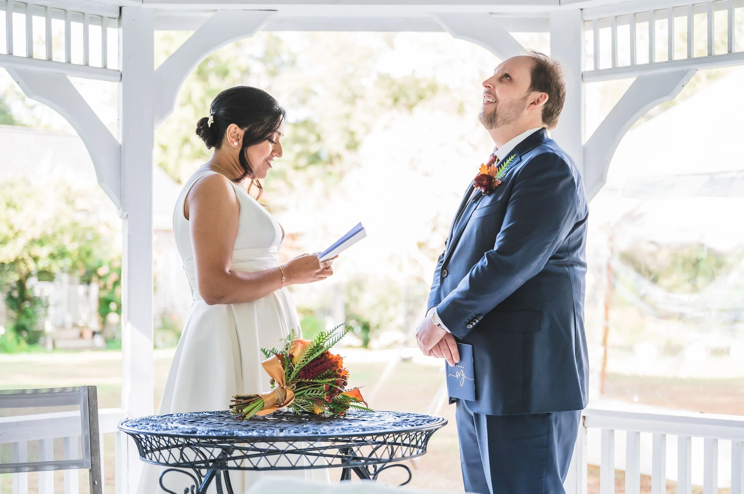 A bride and groom standing under a white pavilion at their wedding ceremony at Lambert's Cove Inn on Martha's Vineyard, with the bride holding a bouquet of flowers and the groom wearing a suit with a boutonniere, looking at each other happily.