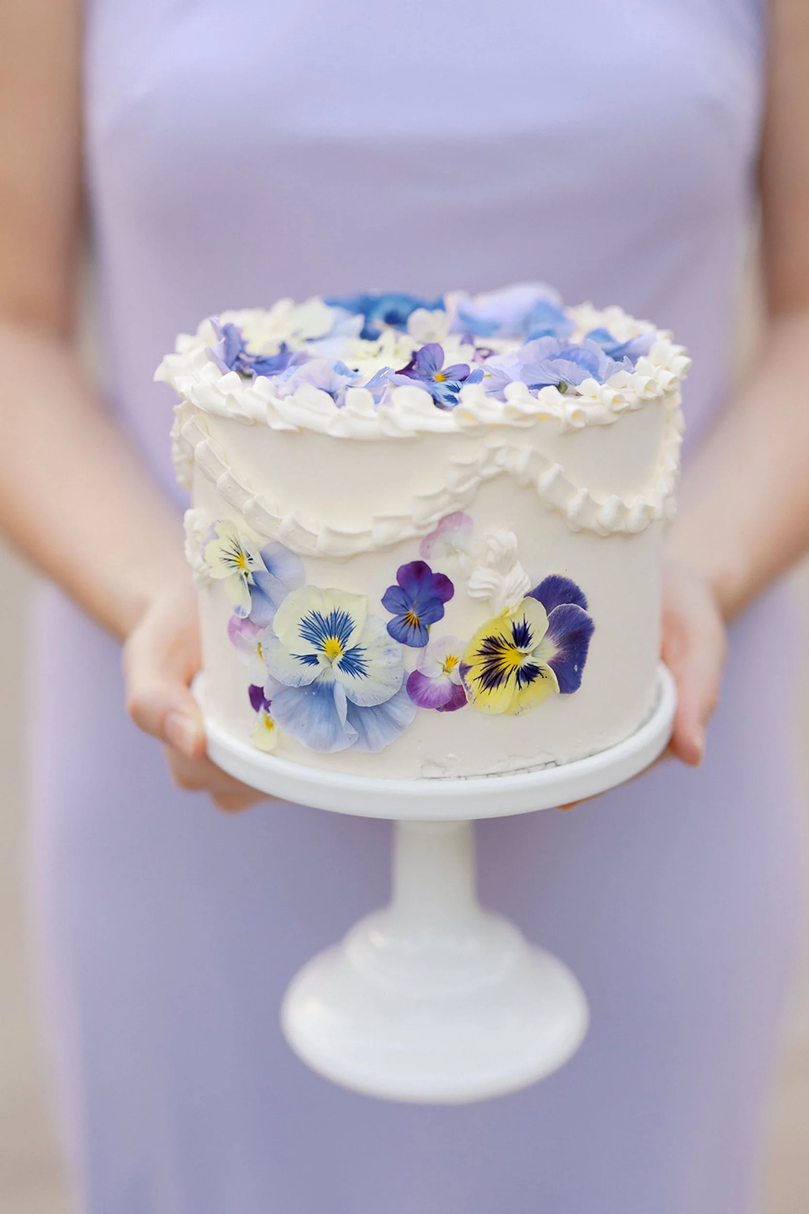 A person in a gray shirt holding a white weding cake decorated with purple and yellow flowers on a white cake stand for wedding ceremony and reception at Lambert's Cove Inn on Martha's Vineyard in West Tisbury, Massachusetts.