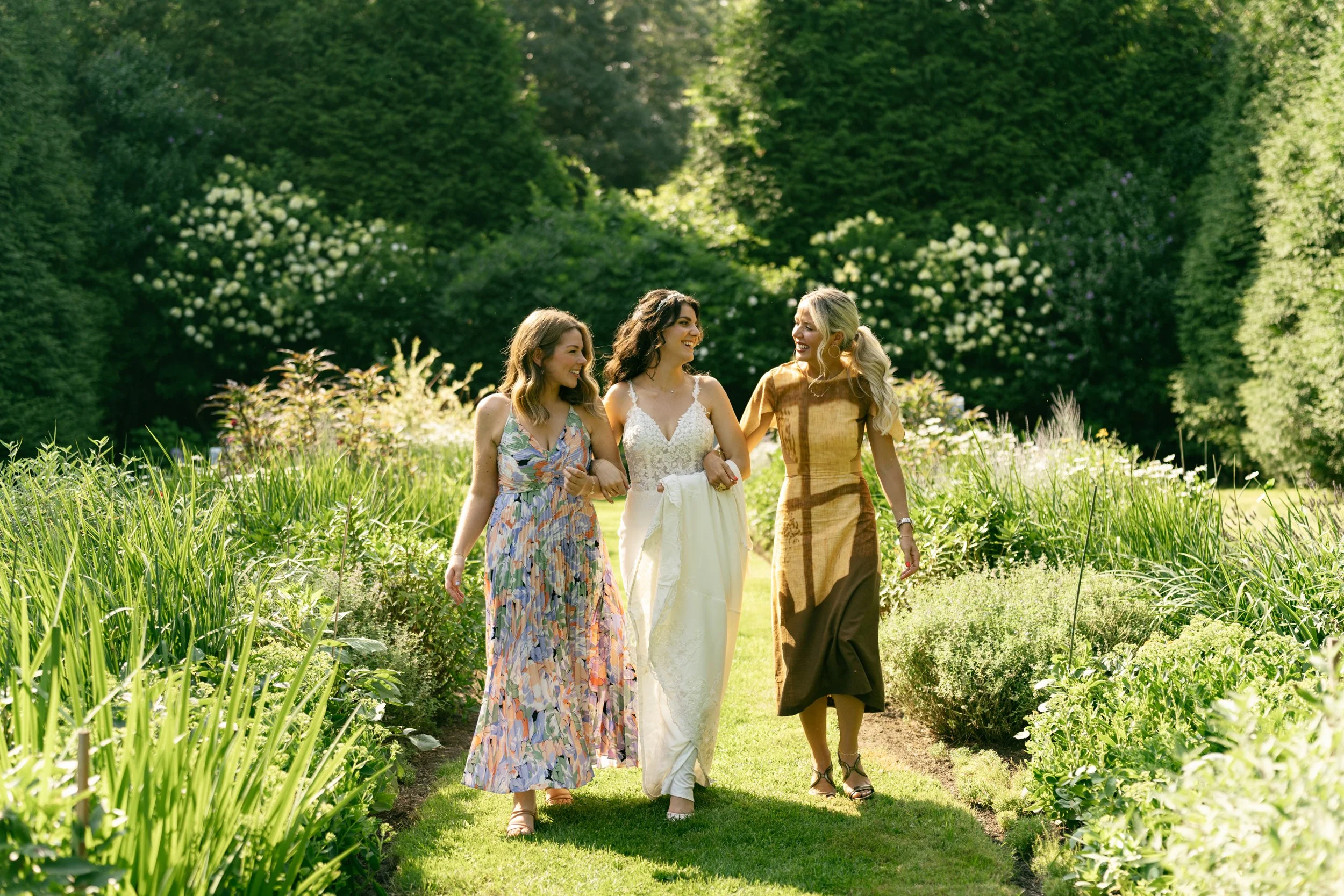 Four women walking together outdoors in the lush English garden at Lambert's Cove Inn on Martha's Vineyard, with one woman in a wedding dress, all smiling and holding hands.