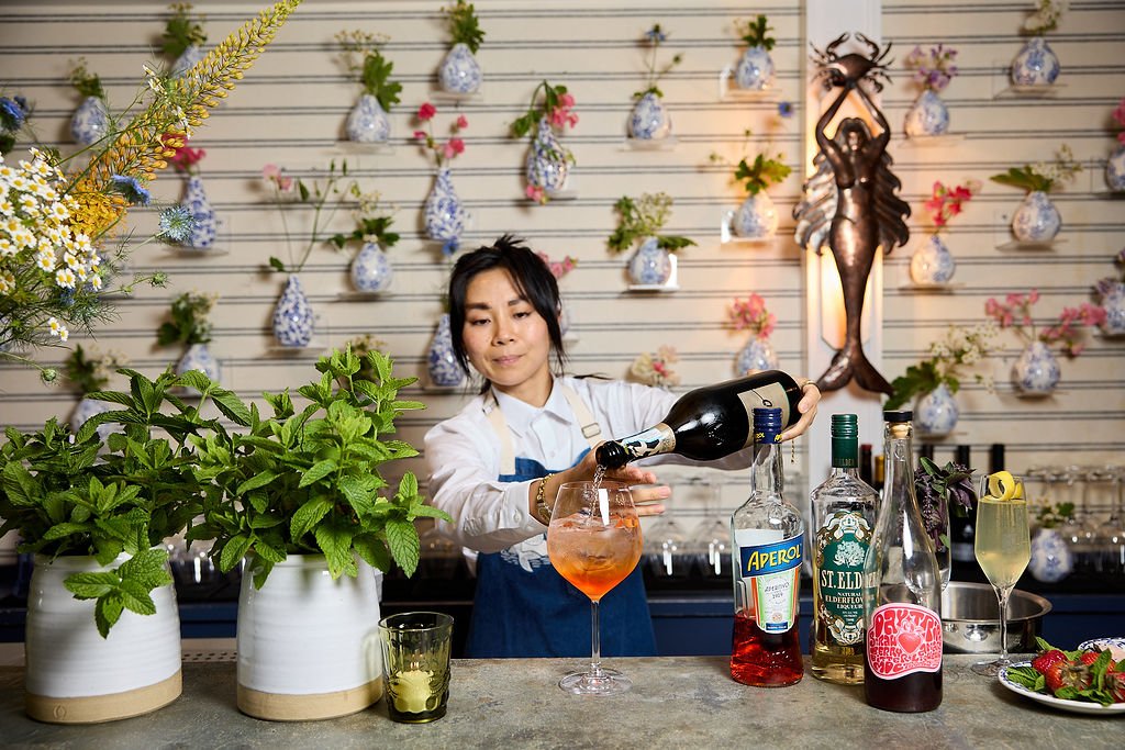A bartender pouring a pink cocktail into a glass in a floral decorated bar with potted mint plants, bottles of alcohol, and a floral arrangement at Mermaid Bar at Lambert's Cove Inn on Martha's Vineyard.