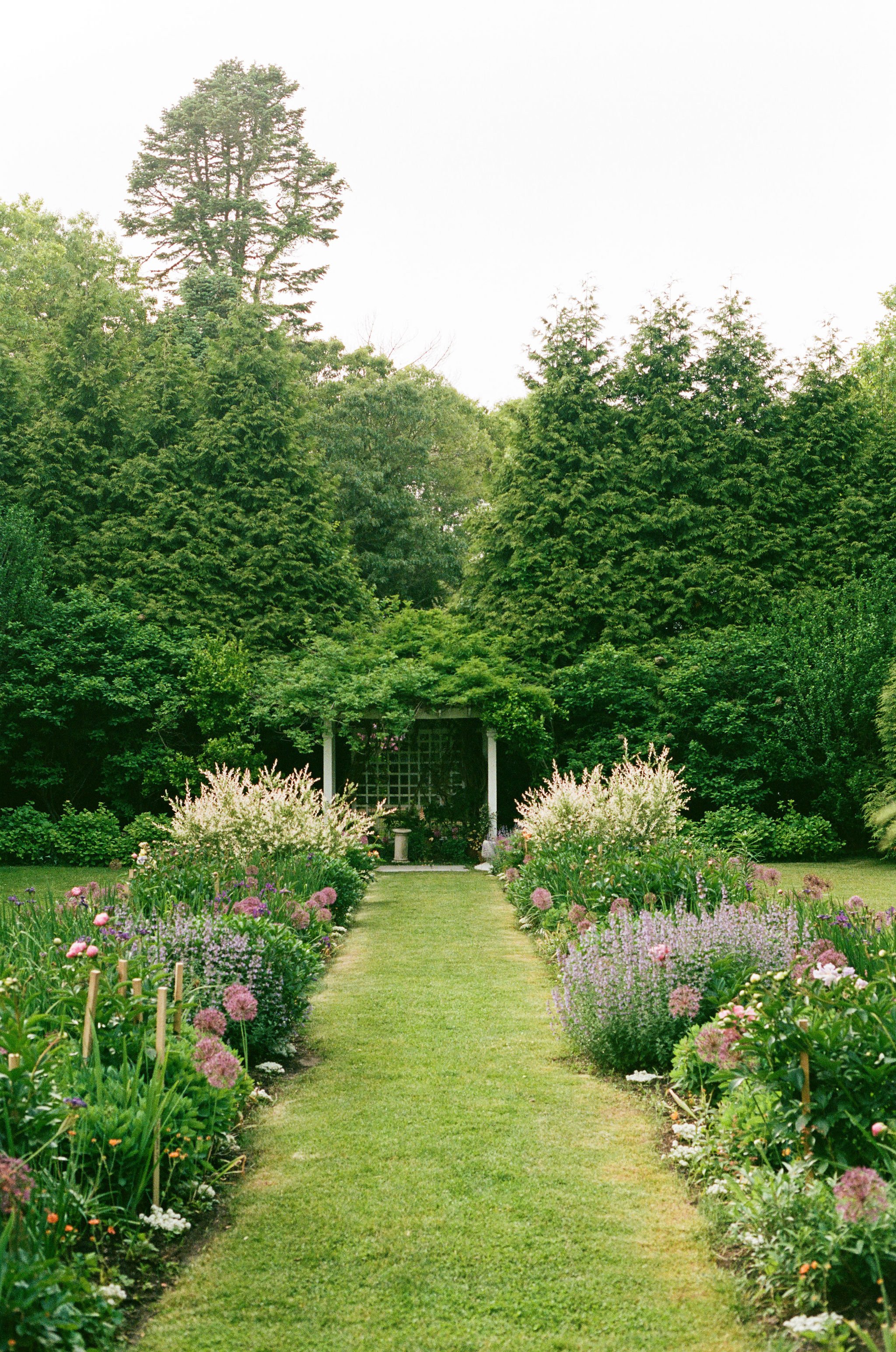A lush garden at Lambert's Cove Inn on Martha's Vineyard with a grassy path, surrounded by various blooming pink, purple, and white flowers, leading to a white pergola or gazebo partially visible in the background, with tall green trees and bushes.