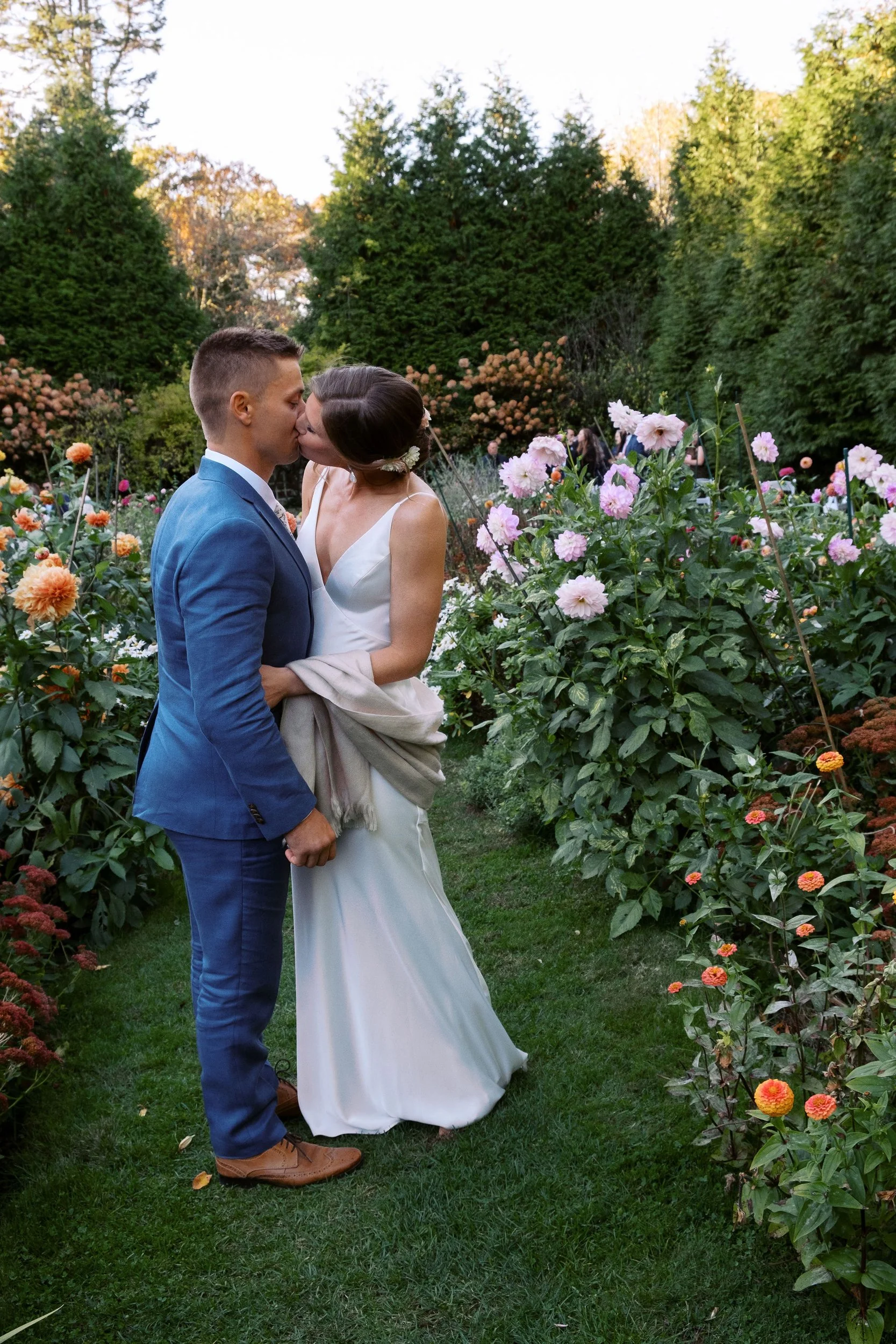 A newlywed couple sharing a kiss in a lush garden filled with vibrant flowers, with trees in the background during the daytime during a wedding at Lambert's Cove Inn on Martha's Vineyard.