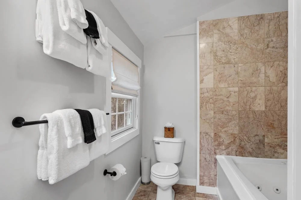 A small bathroom with white walls, a window with a white blind, a toilet, a small basket on top of the toilet, a bathtub with a tan marble tile wall, and laundry towels hanging on black towel bars.