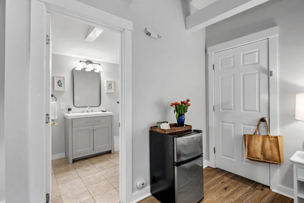 Interior of a small living space with a view into a bathroom, featuring a mini fridge with a bouquet of red tulips and a brown tote bag hanging on a door.