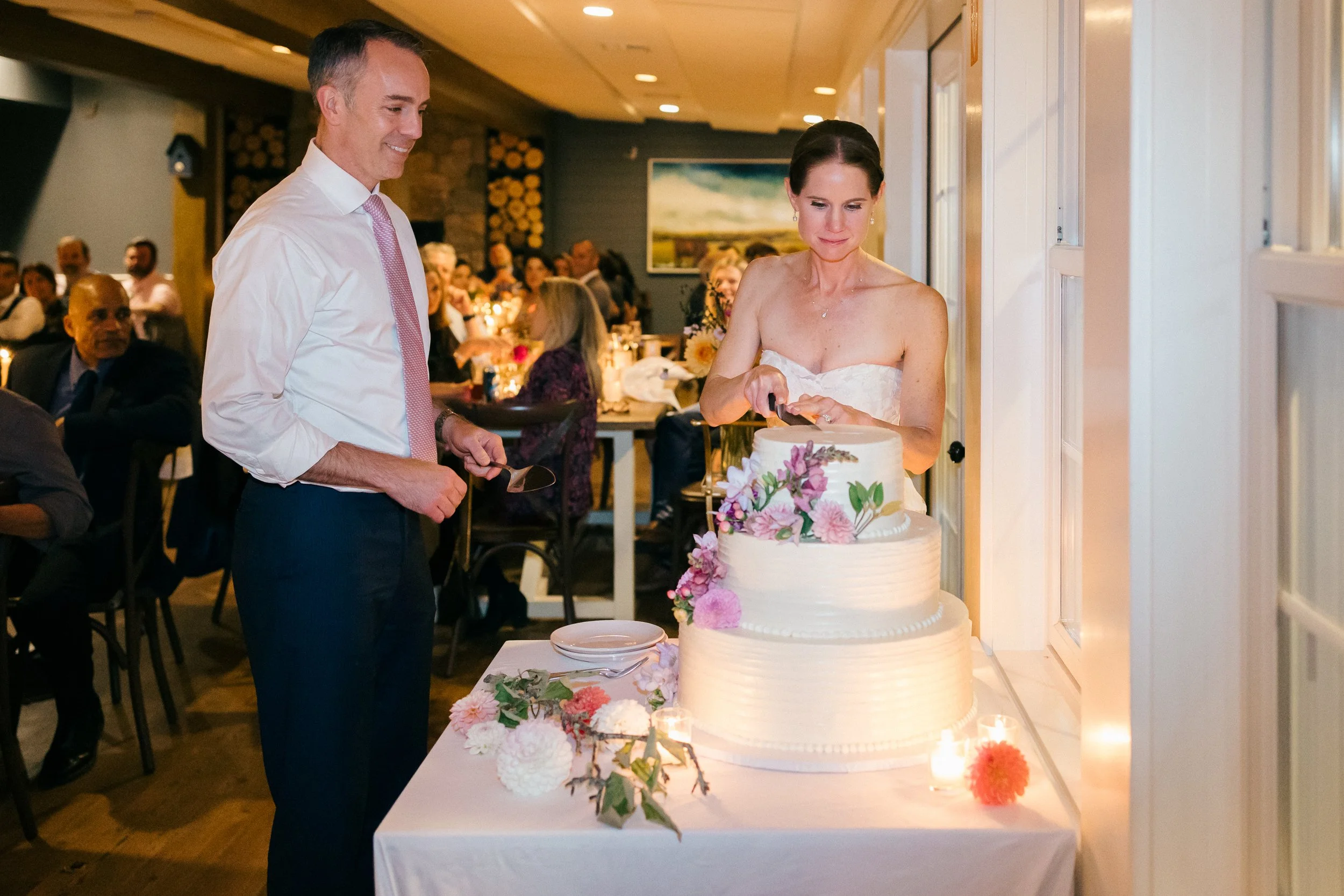 A couple in wedding attire cutting a tiered wedding cake decorated with pink and purple flowers at a wedding reception, with guests seated in the background during a wedding at Lambert's Cove Inn on Martha's Vineyard.