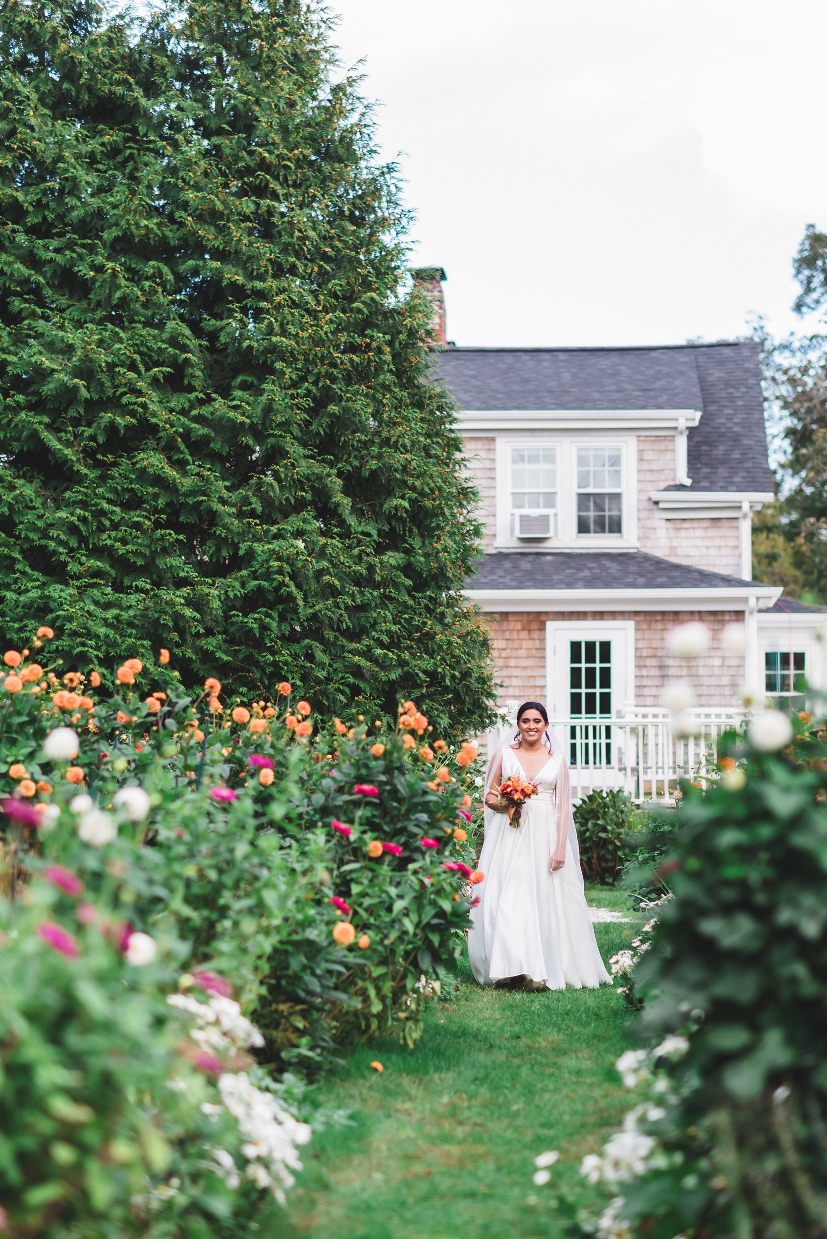 A woman in a wedding dress holding a bouquet, standing in a garden at Lambert's Cove Inn on Martha's Vineyard surrounded by colorful flowers.