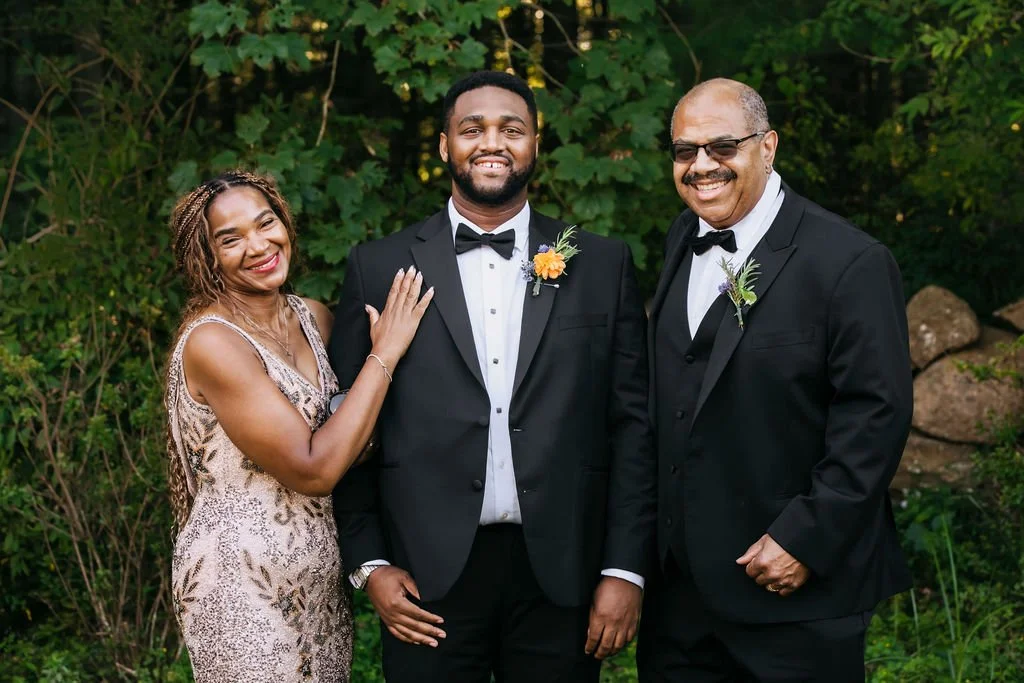 Three people dressed in formal wedding attire standing outdoors in front of greenery, smiling at the camera during a wedding reception at Lambert's Cove Inn on Martha's Vineyard.
