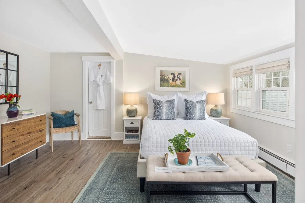 A bright, tidy bedroom with a white bed, two nightstands with lamps, a wooden dresser, a chair, and a window with blinds.
