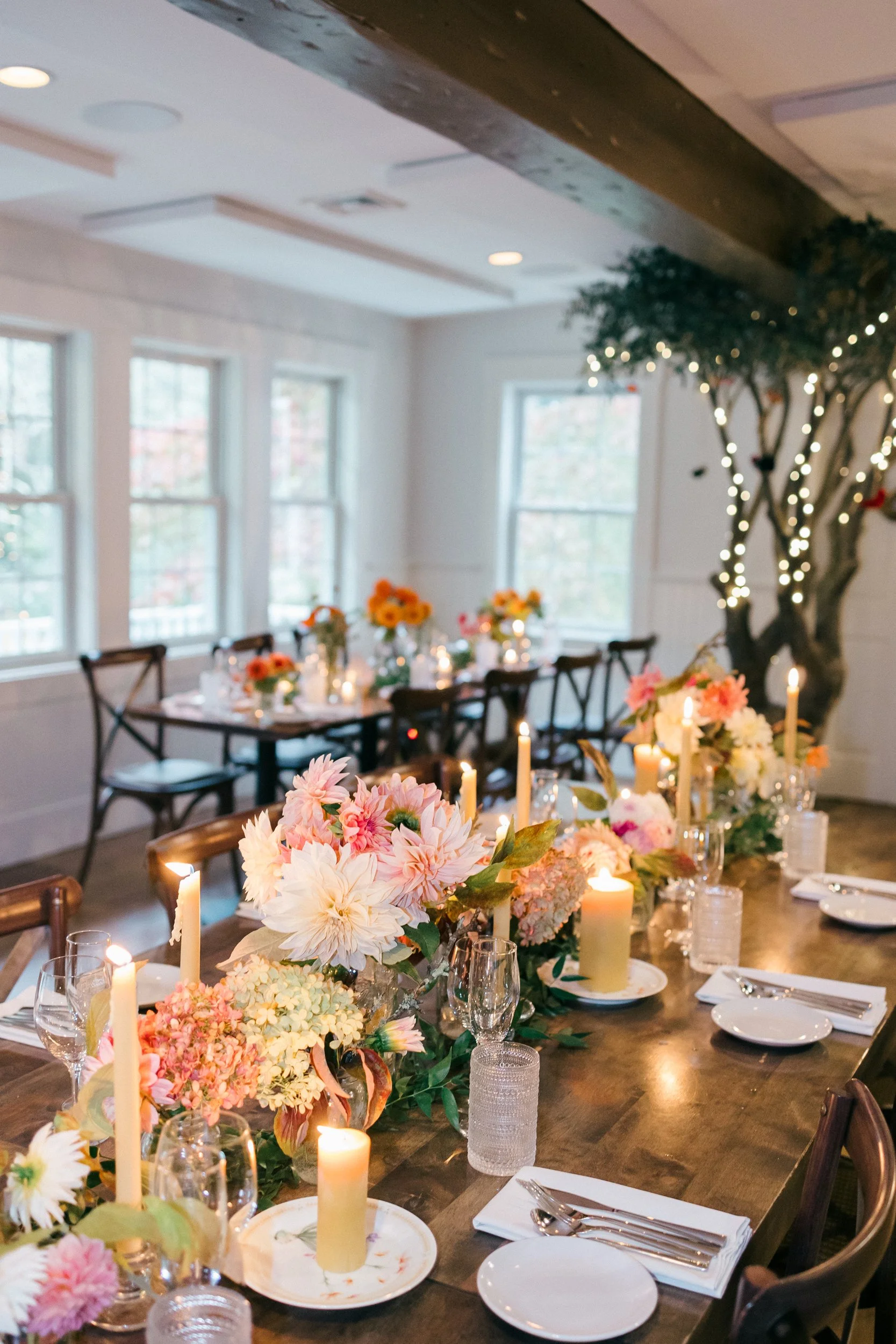 A beautifully decorated indoor dining table with floral arrangements, candles, and place settings, set in a bright room with large windows and distinctive wooden beams on the ceiling at Lambert's Cove Inn on Martha's Vineyard.