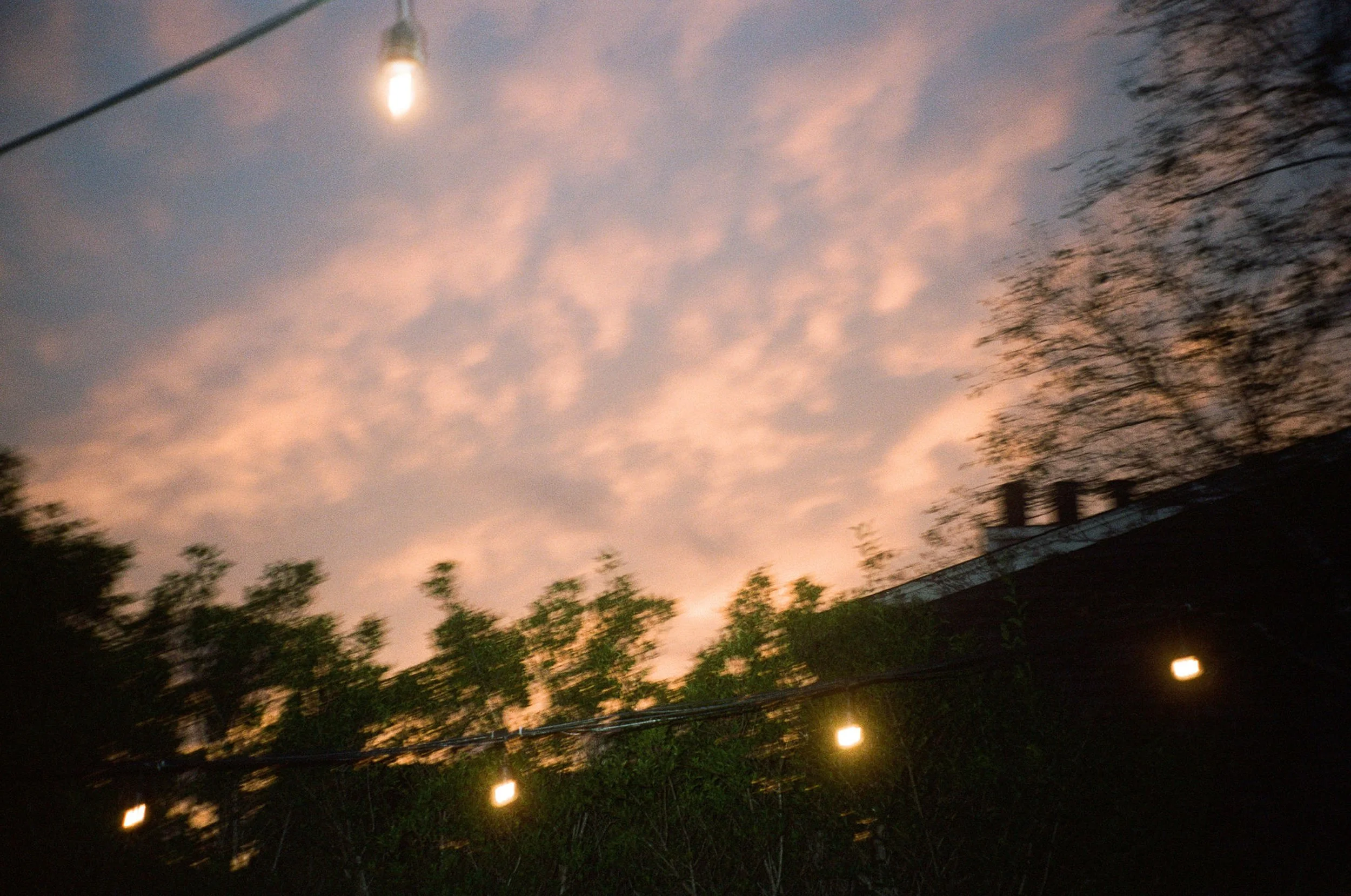 A sunset sky with pink and purple clouds, trees in silhouette, string lights hanging in the foreground, and a rooftop chimney on a building.