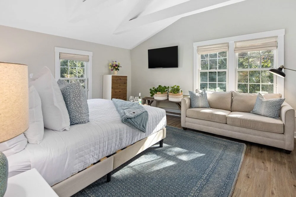 Bright bedroom with a white bed, beige sofa, wooden dresser, and multiple windows letting in natural light.