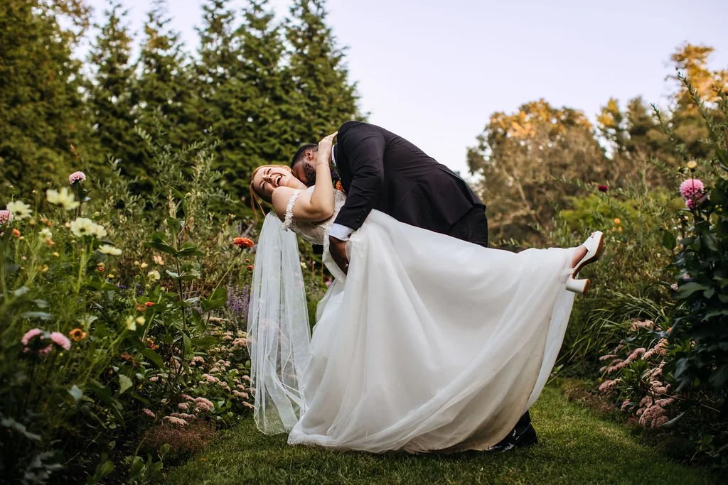 A bride and groom sharing a kiss in a garden with colorful flowers and green trees in the background, the groom dipping the bride, during a wedding ceremony outdoors at Lambert's Cove Inn on Martha's Vineyard.