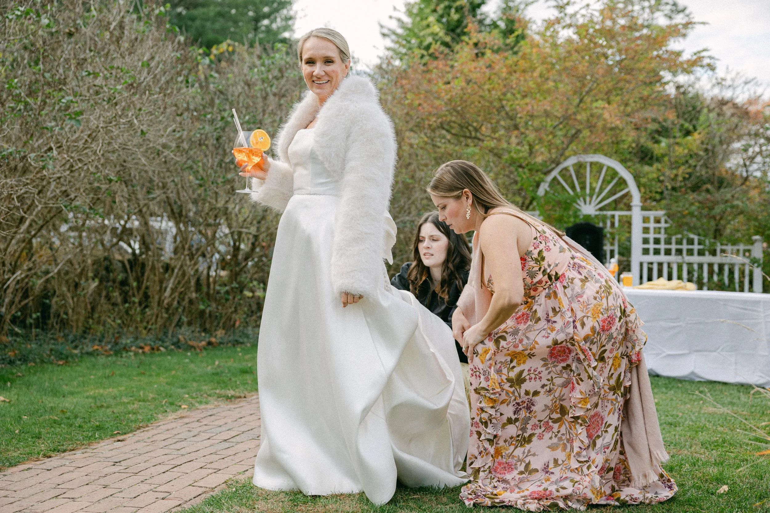 A bride in a white wedding dress and faux fur shawl holds a cocktail glass with a lemon slice, smiling outdoors during daytime. Two women kneel beside her, one in a floral dress, all on a paved walkway with green grass and trees in the background.