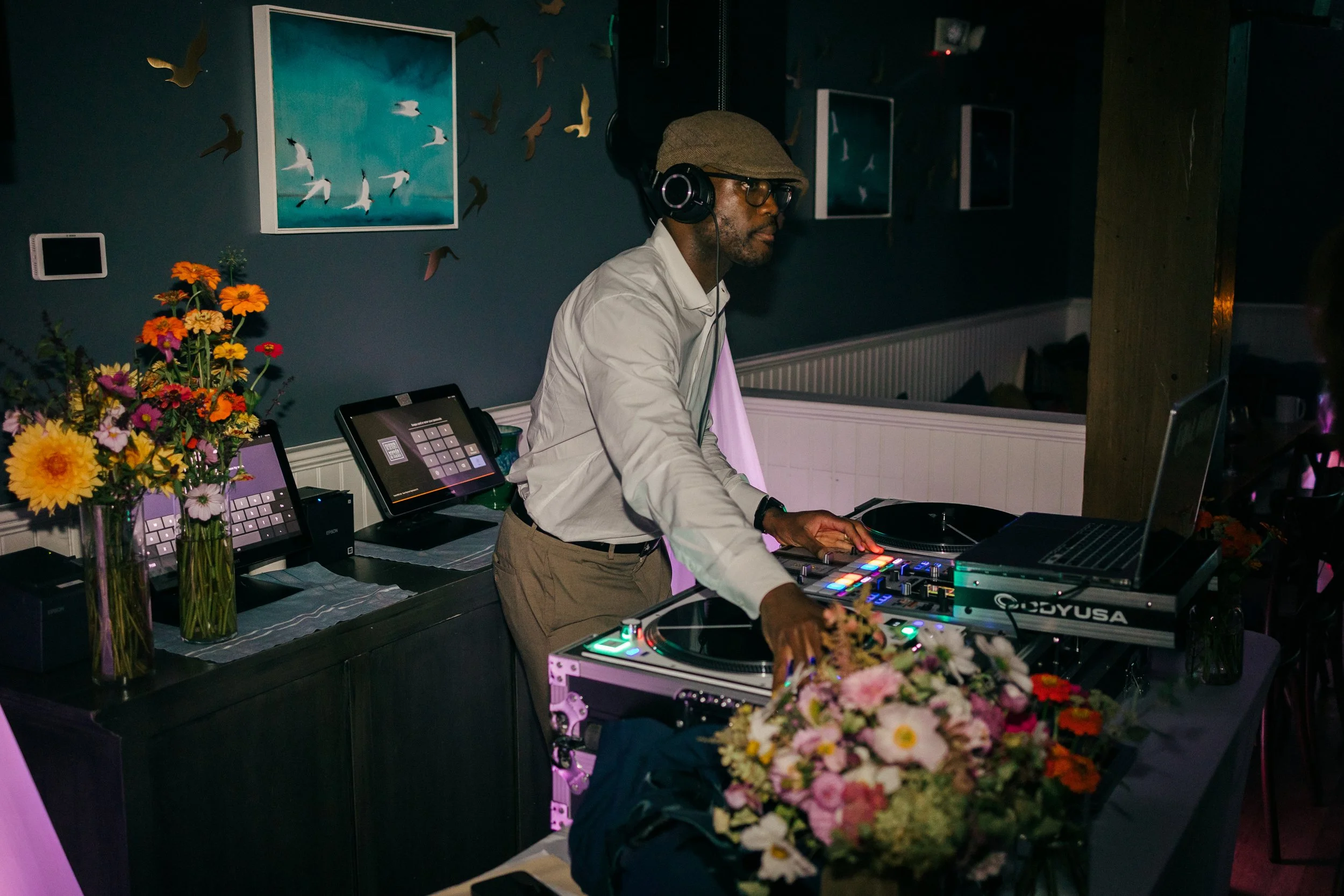 A man DJing at a wedding reception at Lambert's Cove Inn on Martha's Vineyard, wearing headphones, glasses, and a light-colored shirt, using DJ equipment with colorful buttons, surrounded by flowers and framed artwork of seagulls on the wall.