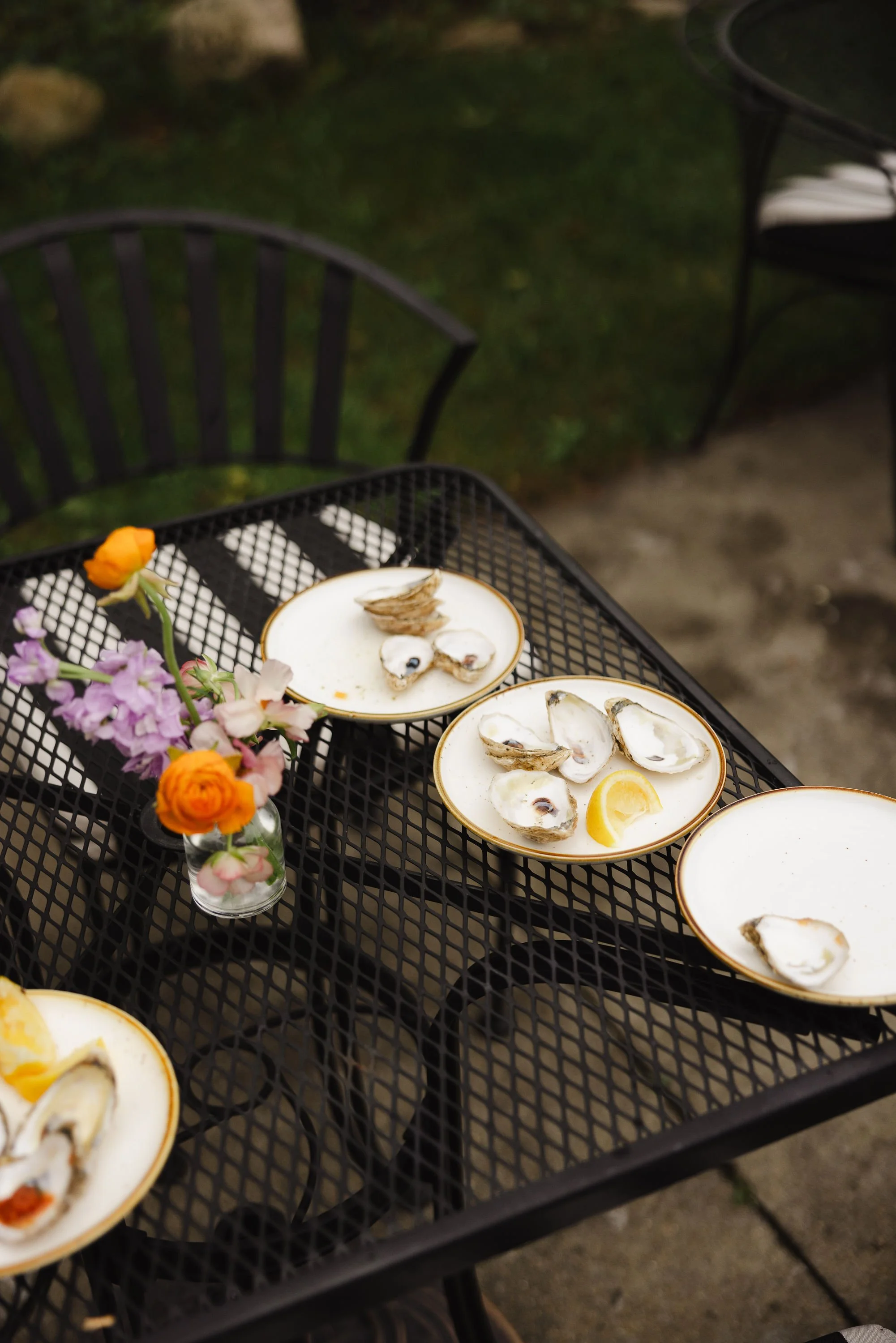Close-up of a black metal outdoor dining table with plates of oysters and lemon wedges, a small bouquet of pink, orange, and purple flowers in a glass vase, and a black chair in the background at Lambert's Cove Inn on Martha's Vineyard.