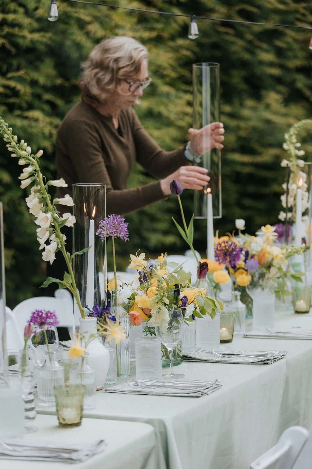 A woman arranging candles and flowers on a table outdoors for a wedding reception at Lambert's Cove Inn on Martha's Vineyard, with greenery in the background.