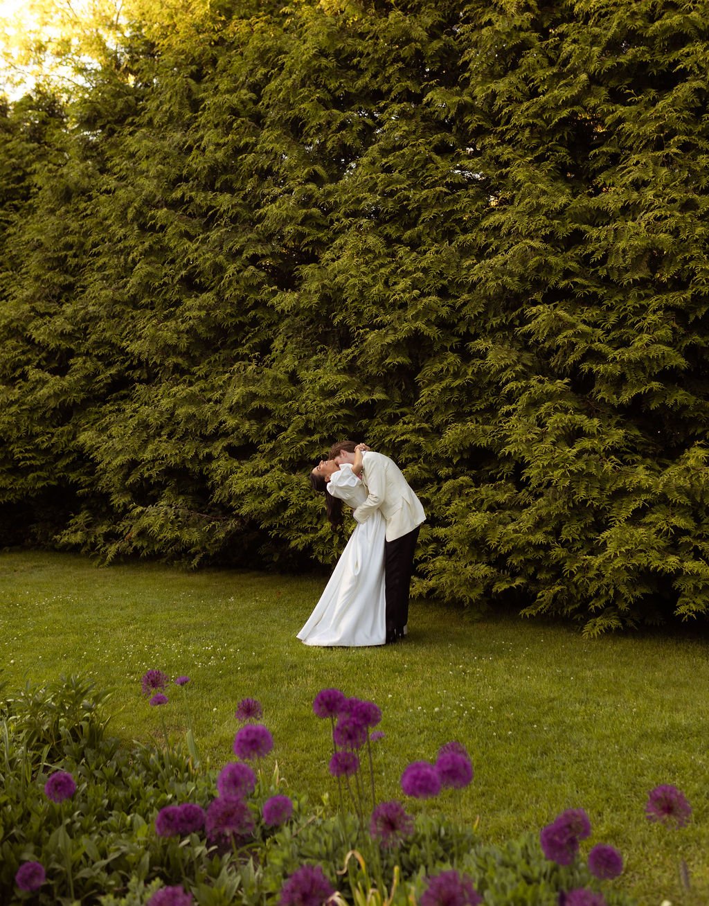 A couple in wedding attire sharing a kiss outdoors next to a large, lush green tree, with purple flowers in the foreground, in the gardens at Lambert's Cove Inn on Martha's Vineyard.
