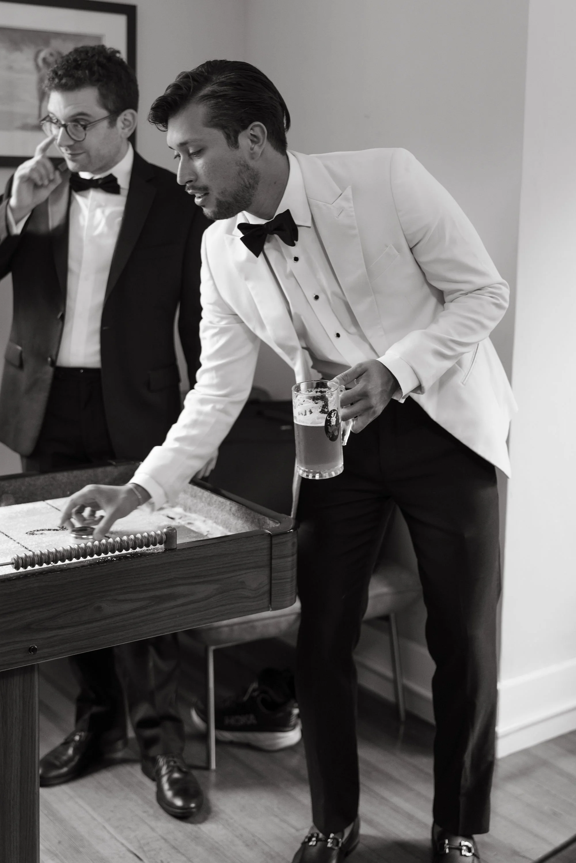 Two men in tuxedos playing a game on a shuffleboard table, one holding a beer mug, in a room with framed art on the wall during a wedding at Lambert's Cove Inn on Martha's Vineyard.