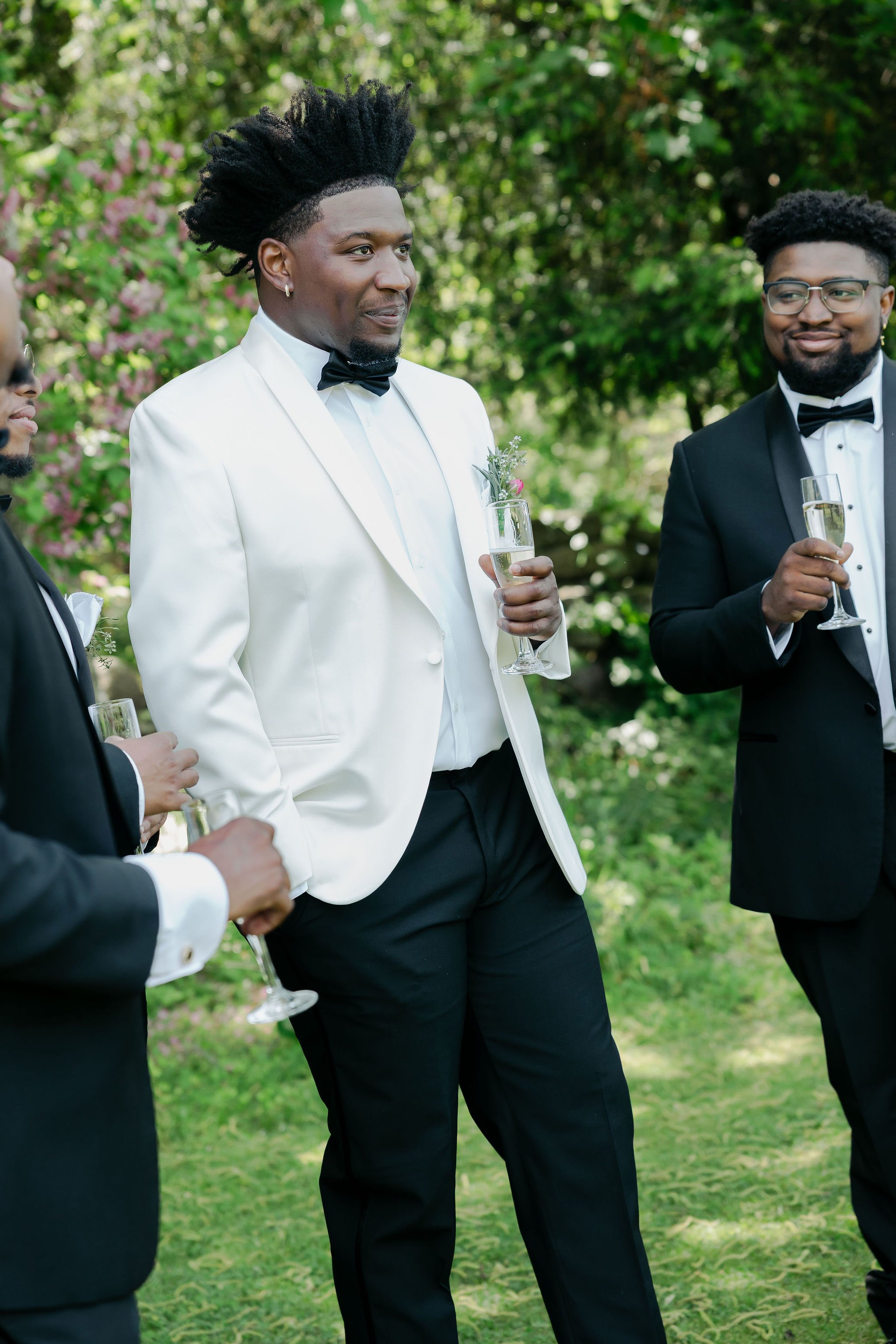 A group of men in tuxedos and bow ties holding champagne glasses, standing outdoors on a lawn with trees and pink flowers in the background during a wedding reception at Lambert's Cove Inn on Martha's Vineyard in West Tisbury.