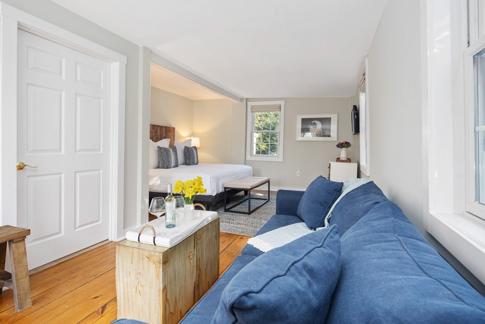 Cozy studio apartment with a blue sofa, a bed with pillows, a nightstand with a lamp, and a window allowing natural light.