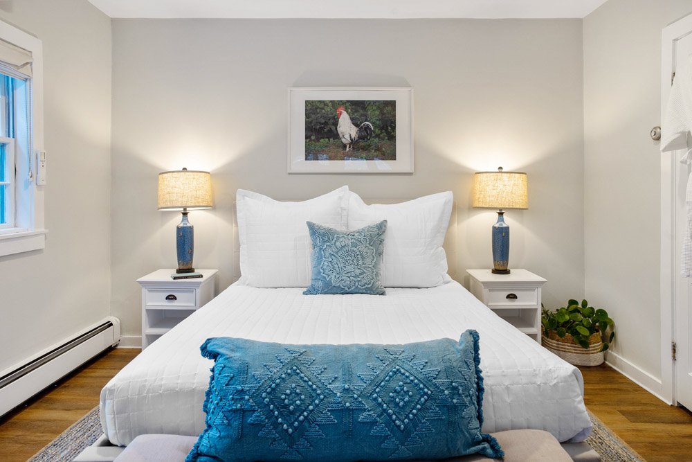 A bedroom with white bedding, two bedside tables with blue lamps, a framed picture of a chicken above the bed, and a blue decorative pillow and throw blanket on the bed.