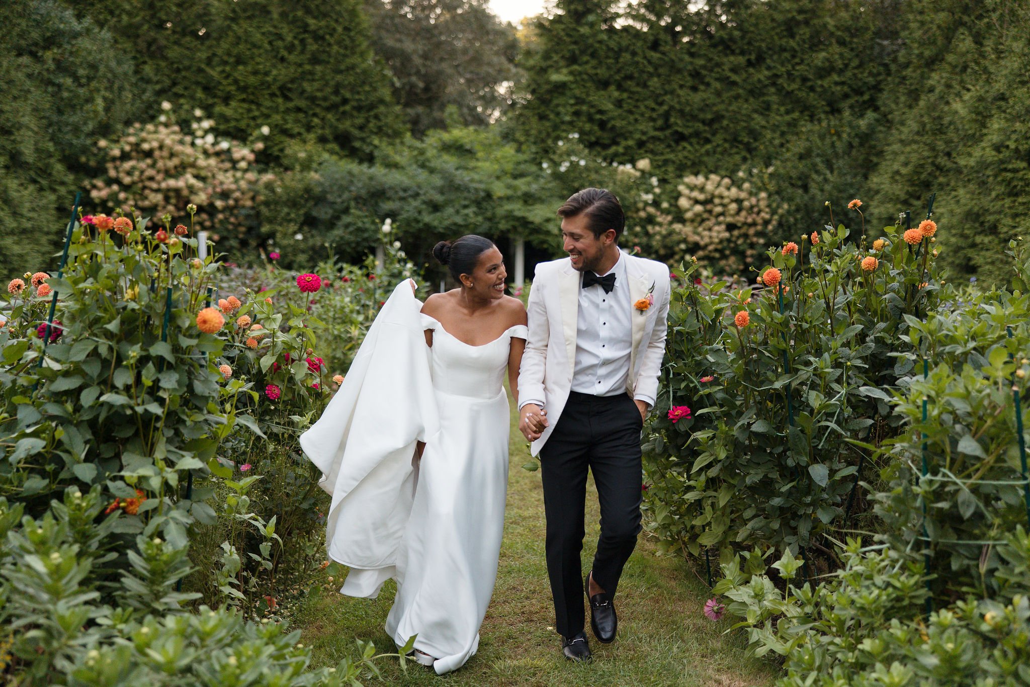 A bride and groom walking hand in hand through a flower garden, smiling at each other, surrounded by colorful blossoms and lush green trees during a wedding at Lambert's Cove Inn on Martha's Vineyard.
