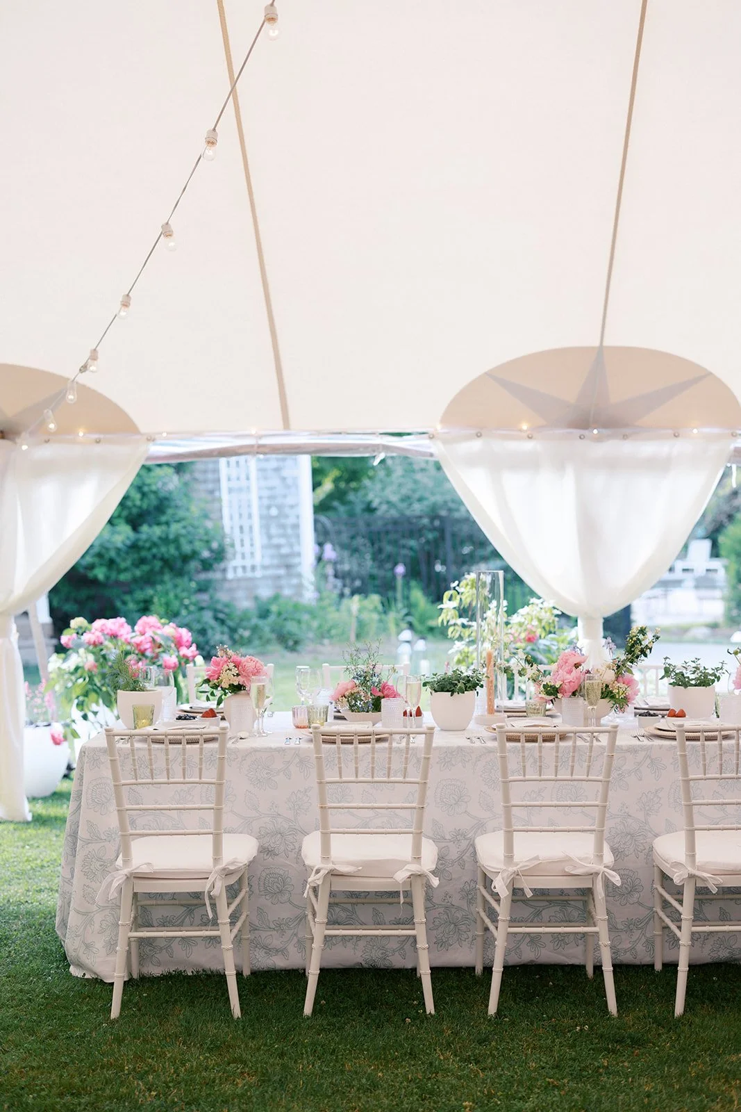 Elegant outdoor dining setup with white tablecloth, pink and white floral centerpieces, white chairs with bows, and a tent with string lights, overlooking a lush garden with trees and a building in the background.