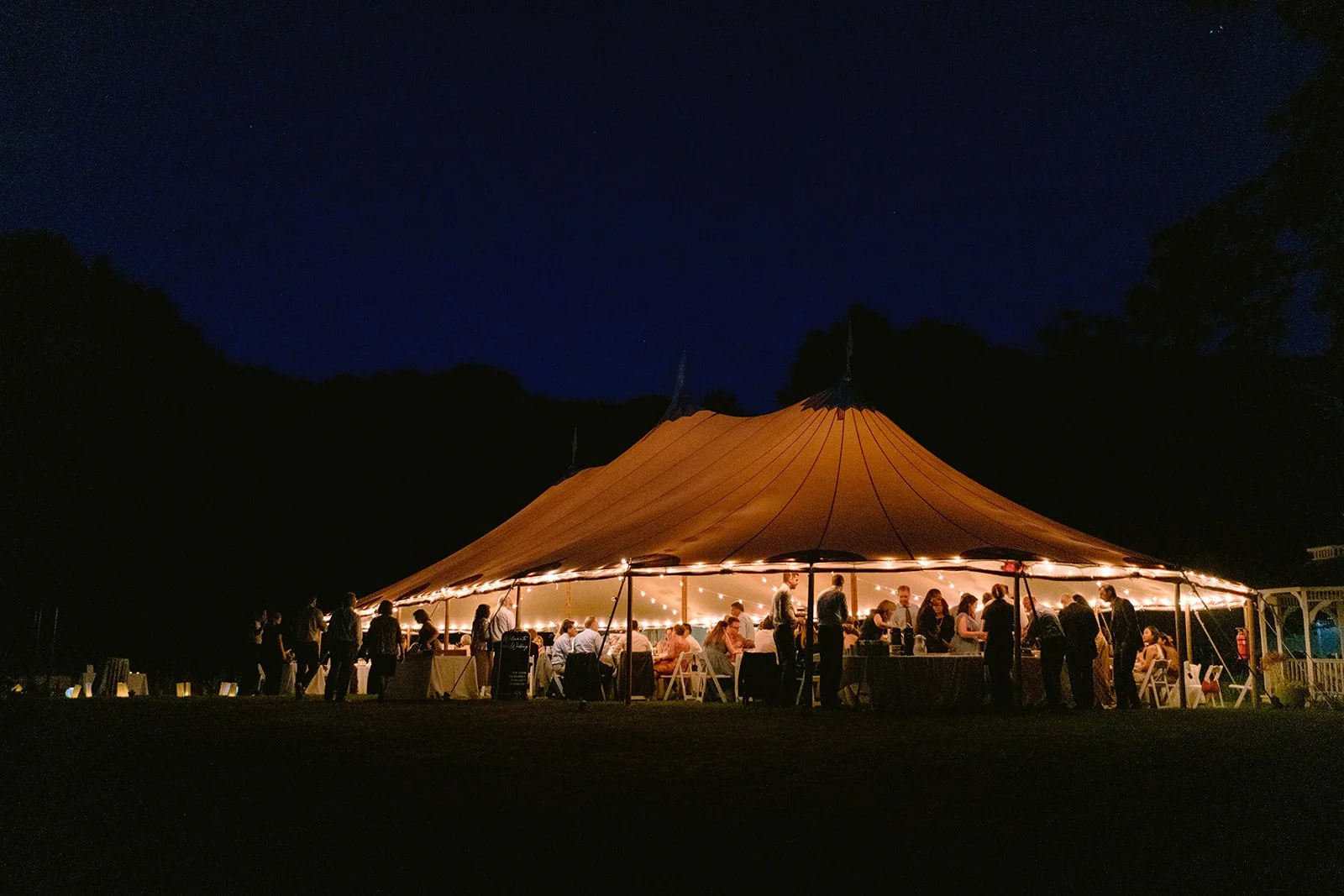 Nighttime wedding reception at Lambert's Cove Inn on Martha's Vineyard under a large orange tent with string lights, with people dining and socializing, surrounded by trees, under a starry sky.