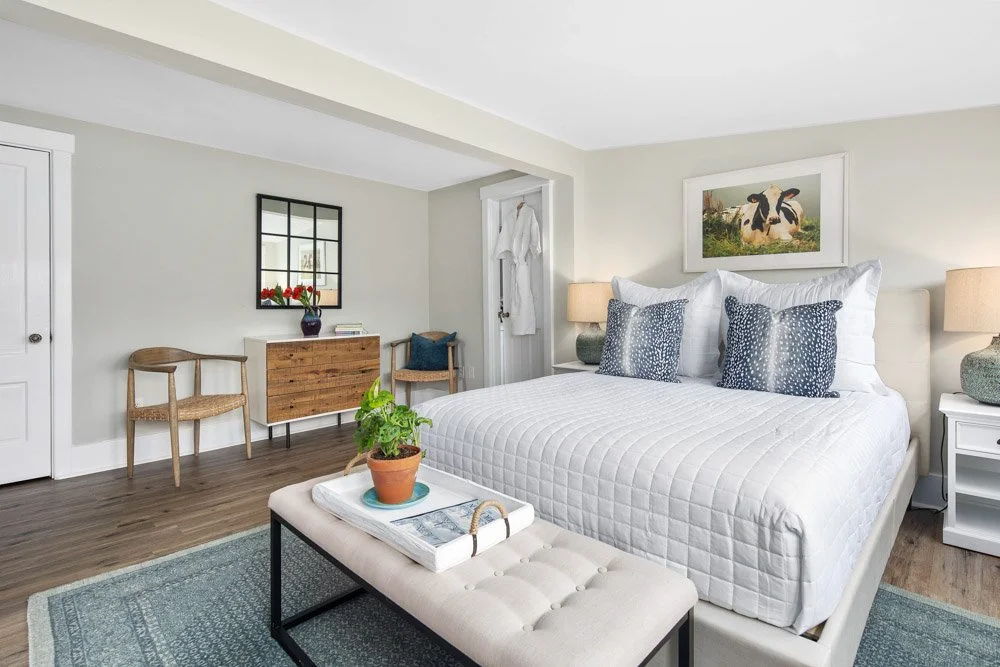A cozy bedroom with a white bed, blue pillows, and bedside lamps, decorated with farmhouse-style artwork and furniture, including a wooden bench with a tray and plant at the foot of the bed.