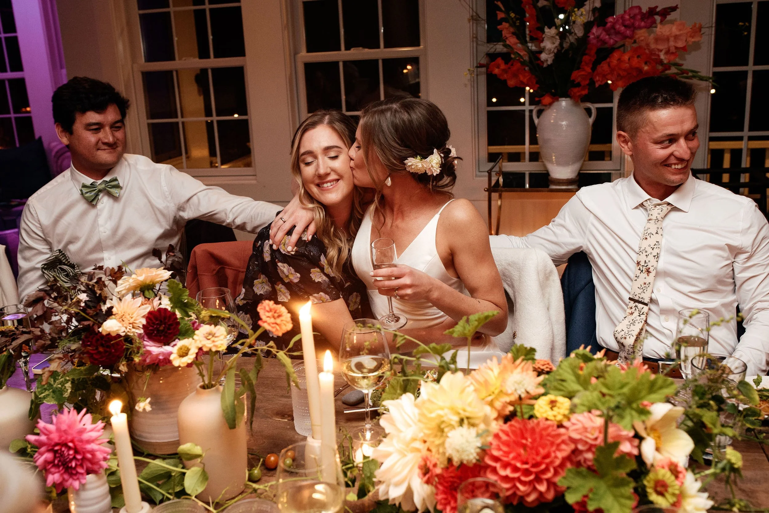 Four people at a table at a celebration, with flowers and candles, one woman kissing another on the cheek, everyone smiling during a wedding at Lambert's Cove Inn on Martha's Vineyard.