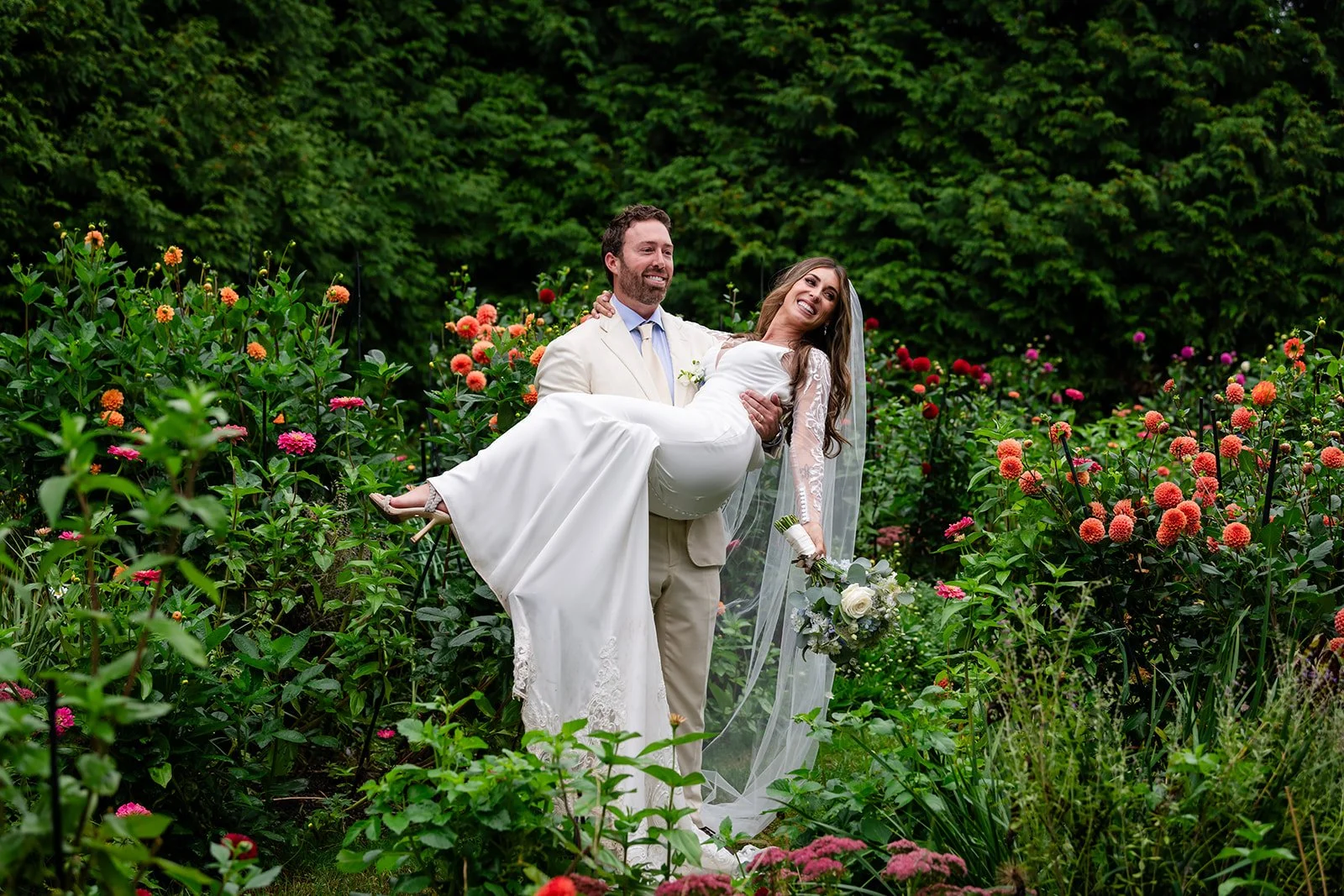 A groom carrying a smiling bride in a garden Lambert's Cove Inn on Martha's Vineyard filled with colorful flowers and lush greenery.