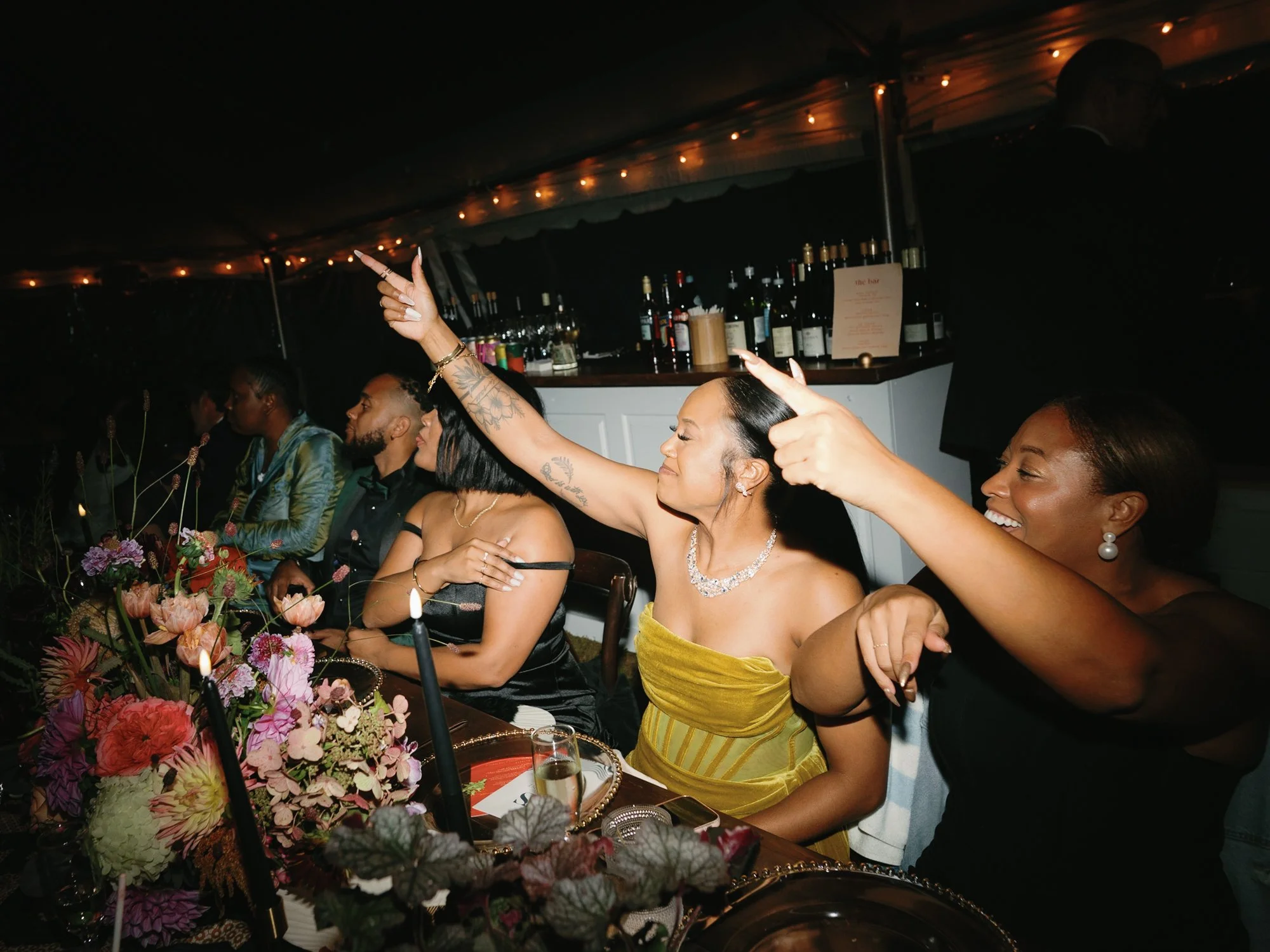 People sitting at a table during a wedding at Lambert's Cove Inn on Martha's Vineyard with floral decorations, candles, and a bar in the background. Women are dressed elegantly, some smiling and gesturing, enjoying the event.