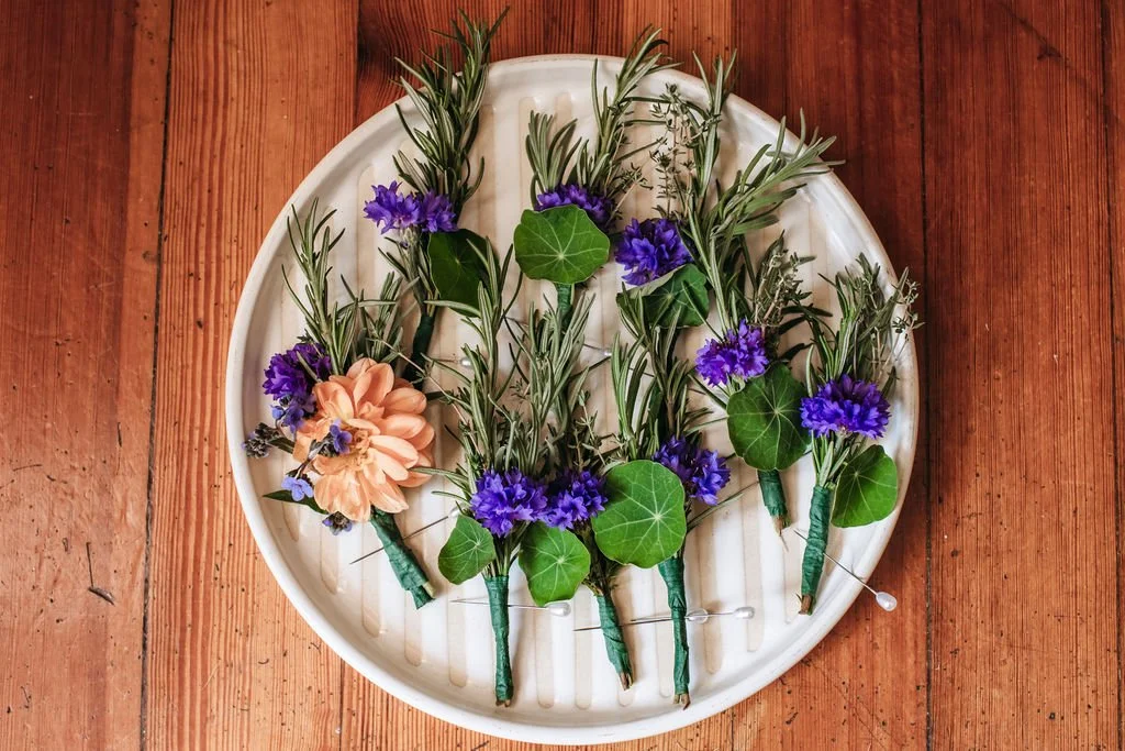Arrangement of boutonnieres with purple, peach, and green flowers on a white oval platter on a wooden table. The flowers were custom grown and designed by Bridget Sampson at Lambert's Cove Inn on Martha's Vineyard.