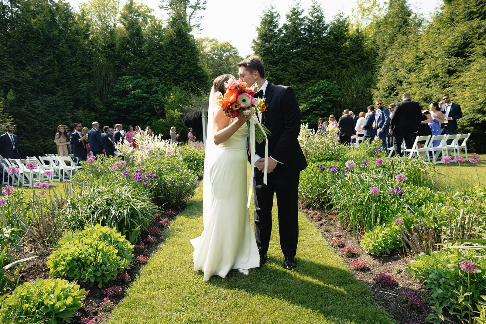 Couple gets married outdoors on a sunny day at Lambert's Cove Inn on Martha's Vineyard, standing on a grassy aisle lined with colorful flowers, with guests in formal attire in the background.