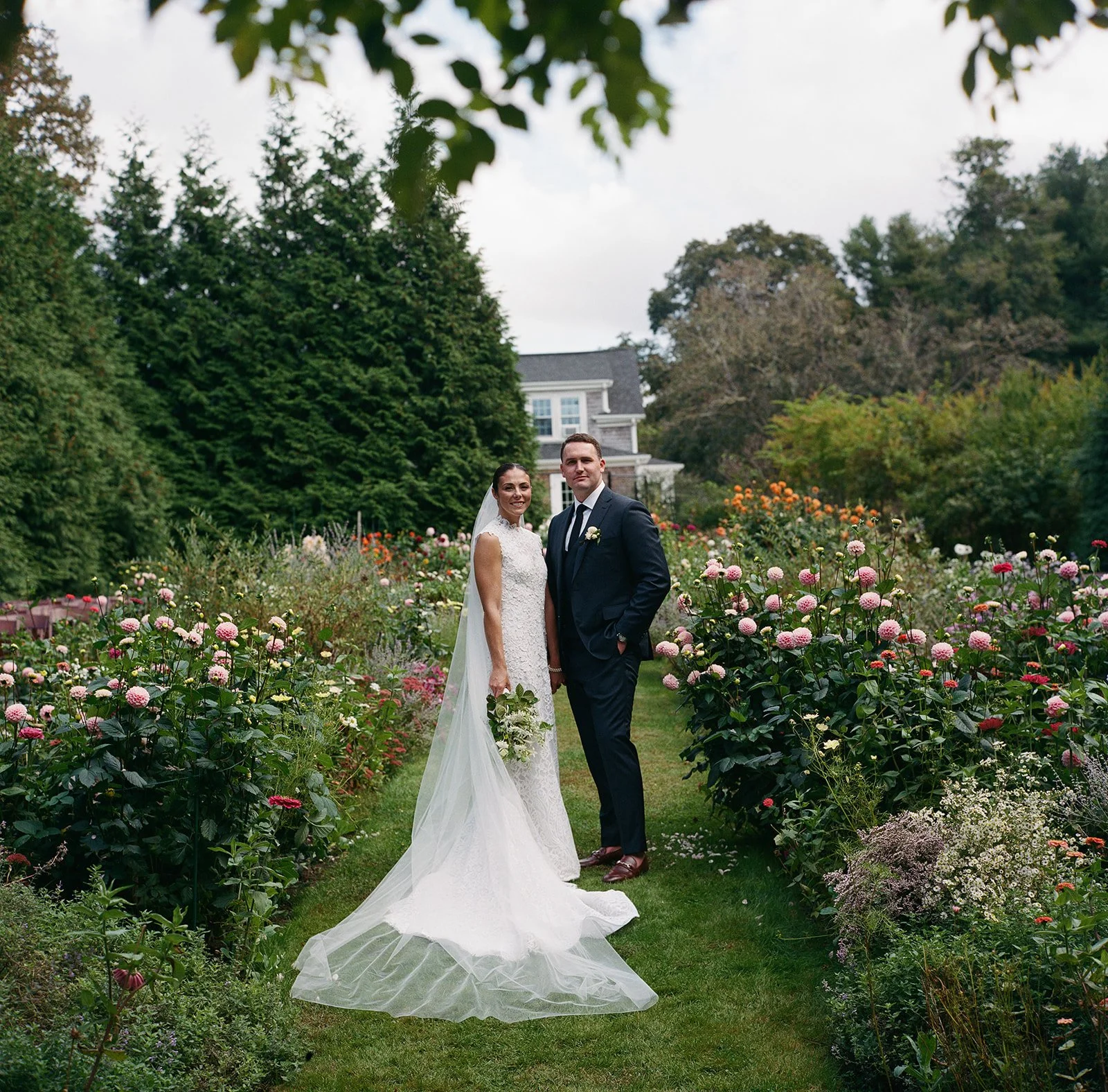 A bride and groom standing together in a garden filled with pink, white, and purple flowers, with a house and trees in the background during a wedding reception at Lambert's Cove Inn on Martha's Vineyard.