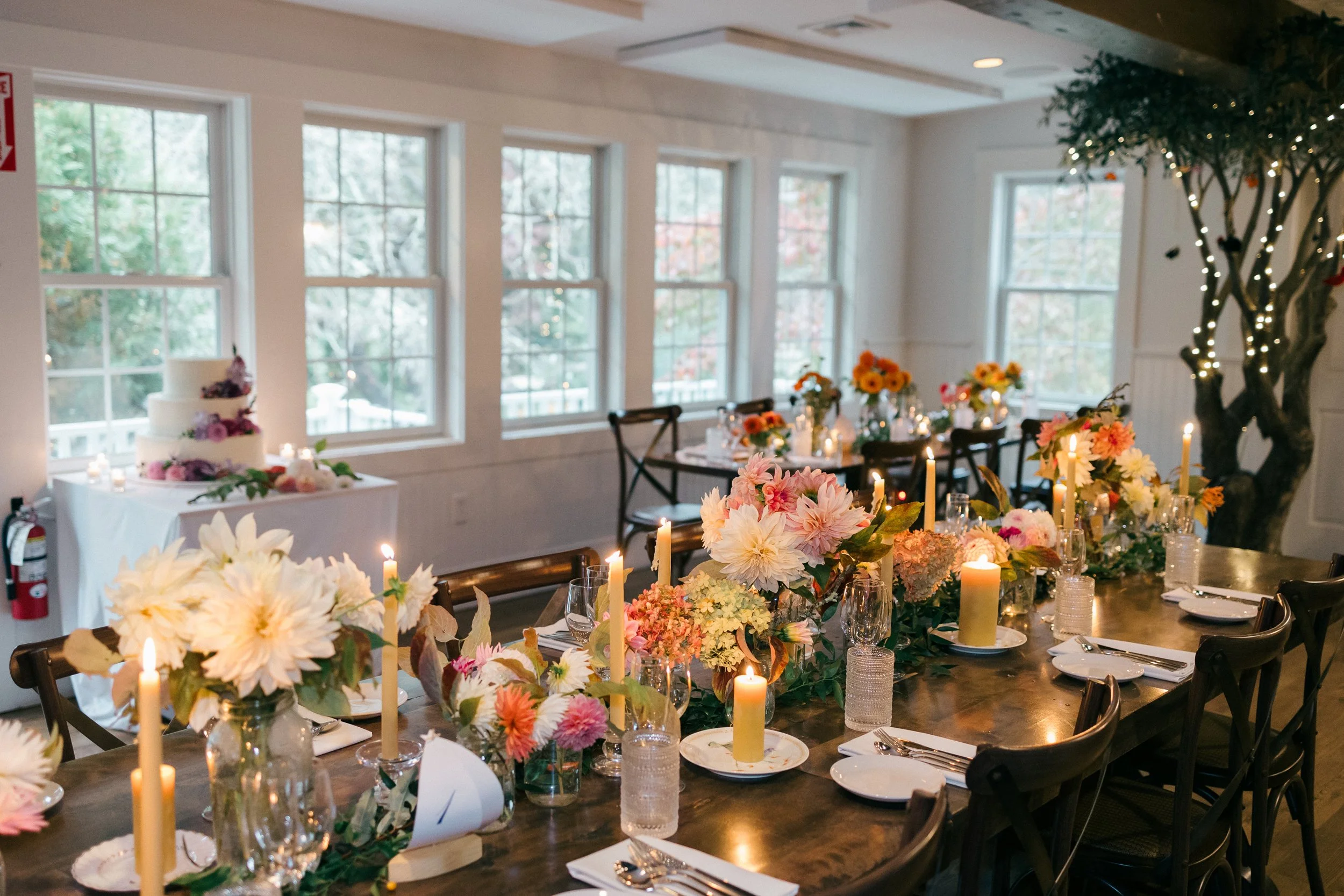 Interior of a decorated event space at Lambert's Cove Inn on Martha's Vineyard with large windows, floral centerpieces, candles, and a wedding cake on a table in the background.