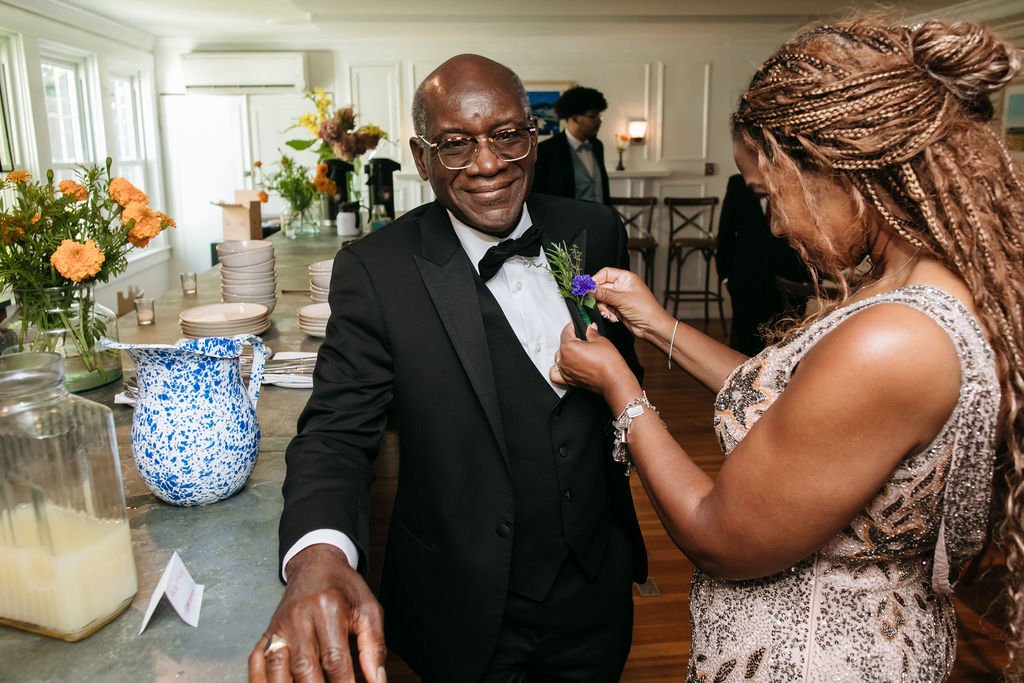 A man in a tuxedo having a boutonniere pinned on his lapel by a woman in a silver dress at a formal event or wedding reception, with a decorated table and floral arrangements in the background at Lambert's Cove Inn on Martha's Vineyard.