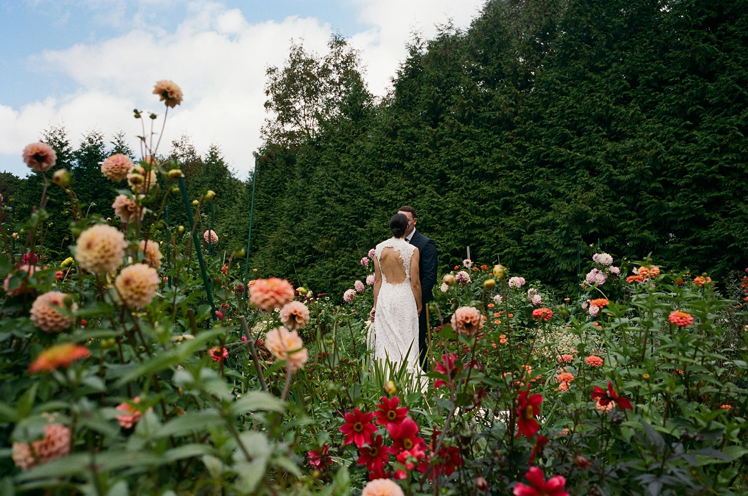 A bride and groom standing in a garden of colorful flowers, with lush green trees and a partly cloudy sky in the background.