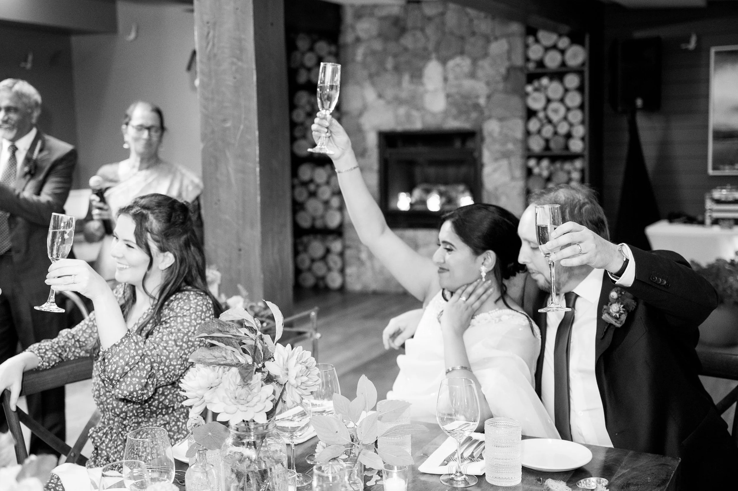 People at a wedding at Lambert's Cove Inn on Martha's Vineyard, sitting at a decorated table with glassware and flowers, raising glasses in a toast, in a cozy indoor setting with a stone fireplace in the background.