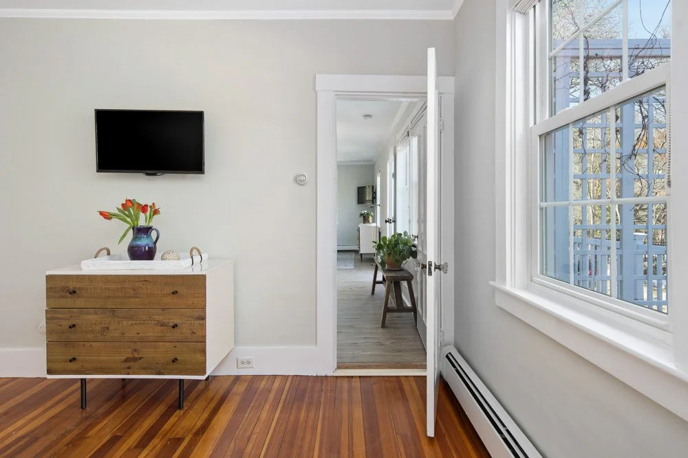 Empty room with wooden floor, white walls, and a wall-mounted TV, with a dresser topped with a vase of tulips. An open door leads to a hall with additional furniture and large windows.