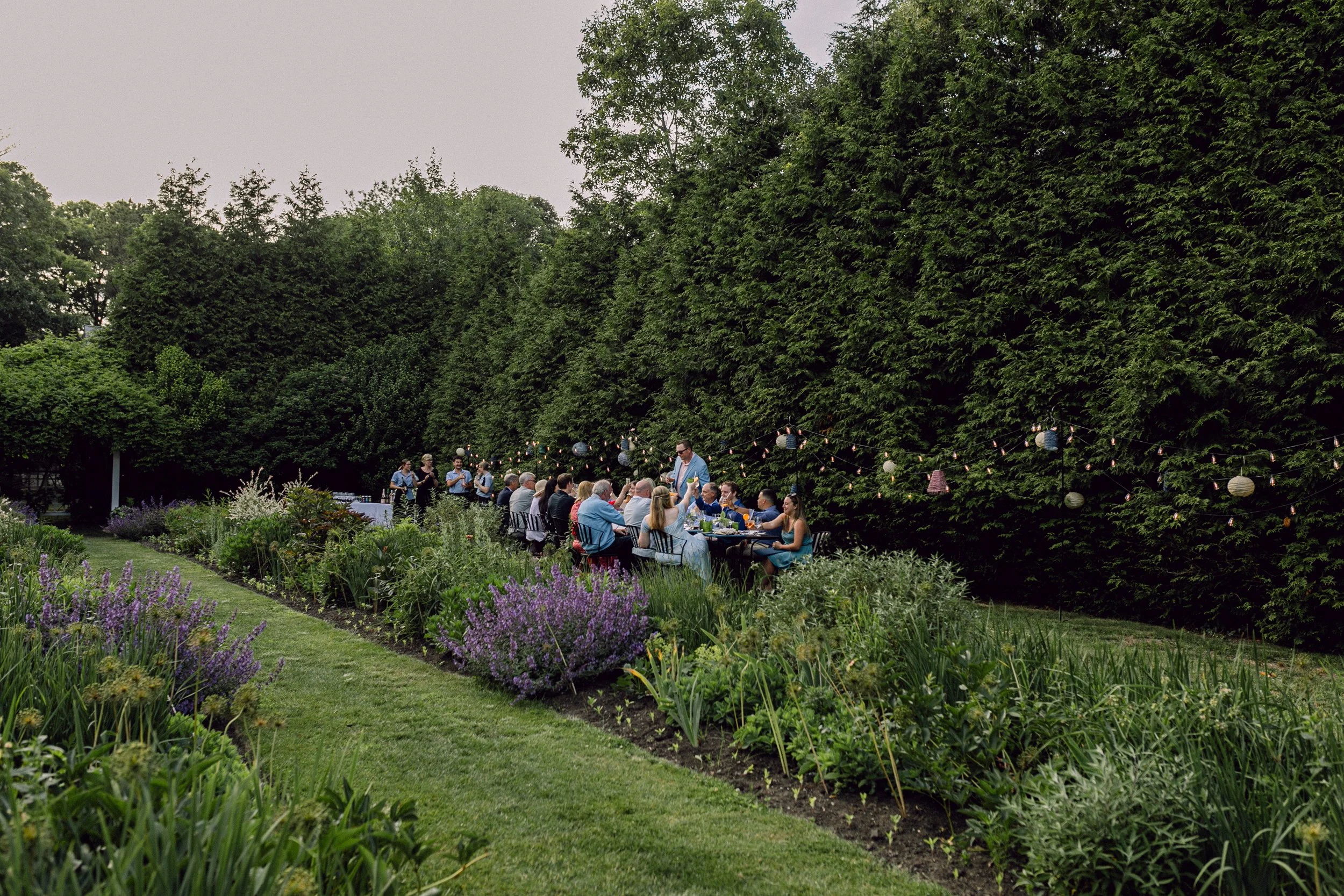 People attending an outdoor wedding reception at Lambert's Cove Inn on Martha's Vineyard in a garden with lush green trees and flowering plants, decorated with string lights and paper lanterns.