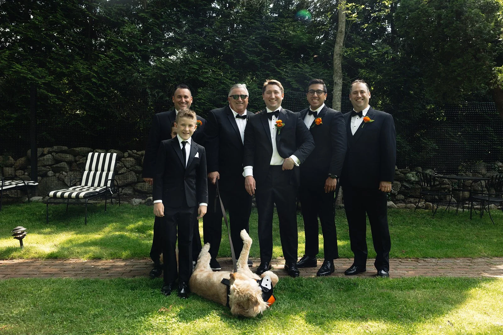 A group of six men in tuxedos with orange boutonnieres and a young boy in a tuxedo standing on a lawn with a dog lying on the grass during a wedding at Lambert's Cove Inn on Martha's Vineyard. There are chairs and a stone wall in the background.