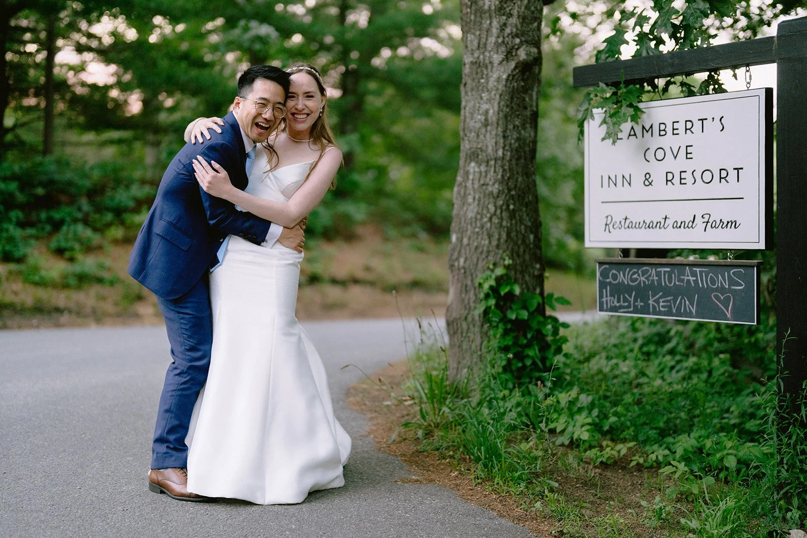 A newlywed couple hugging and laughing outdoors near a sign that reads Lambert's Cove Inn & Resort and a congratulatory message for Holly and Kevin. The setting is lush and green with trees and a paved path.
