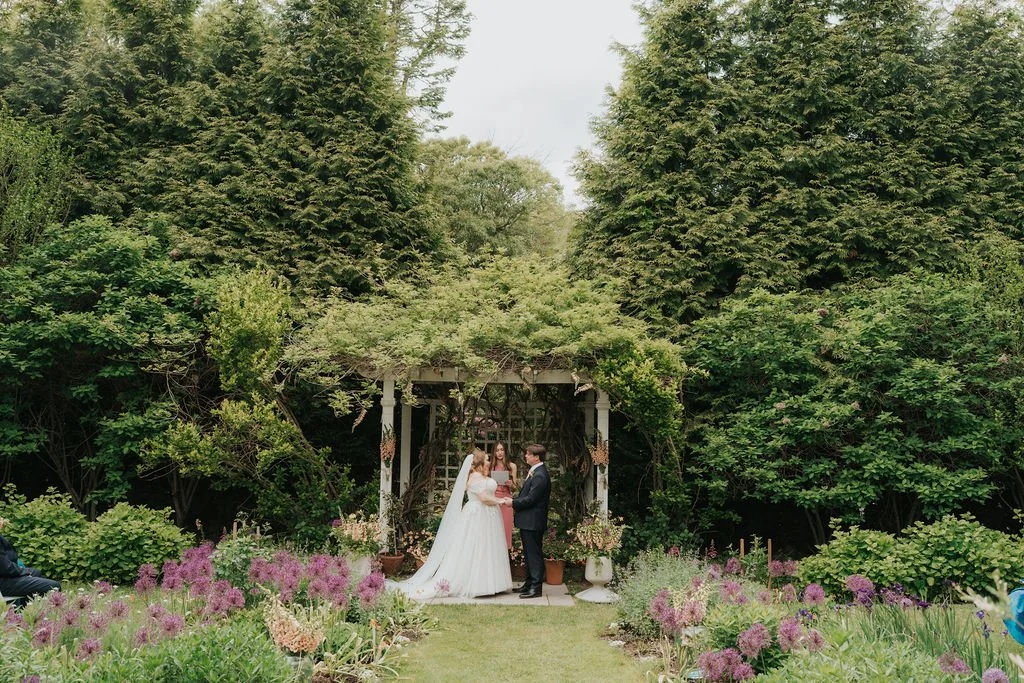 Bride and groom exchanging vows under a floral arch in a lush garden. The wedding is at Lambert's Cove Inn on Martha's Vineyard. 