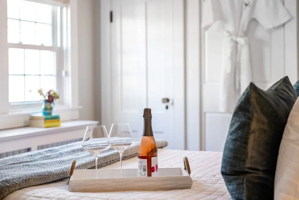 A tray with an unopened bottle of rosé wine and two empty wine glasses on a bed with pillows, in a bright bedroom with a window, a white door, and a vase of flowers on the windowsill.