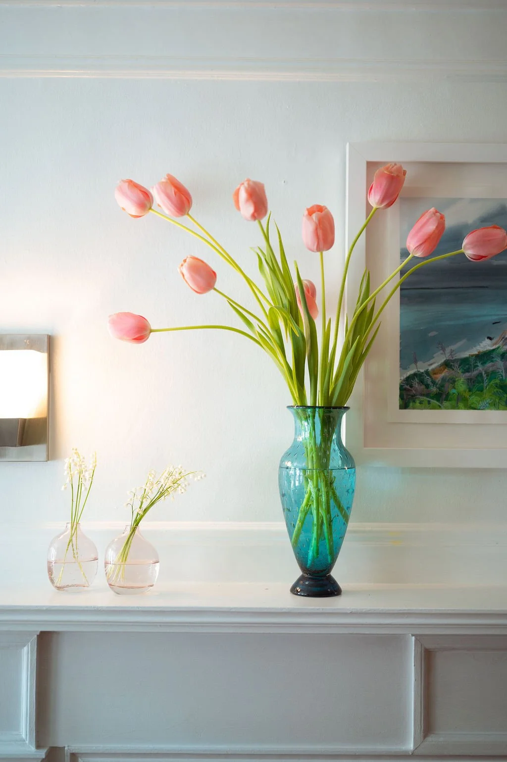 A large blue glass vase with pink tulips on a white mantel, with two smaller pink vases and a framed landscape painting on a white wall at Lambert's Cove Inn on Martha's Vineyard.