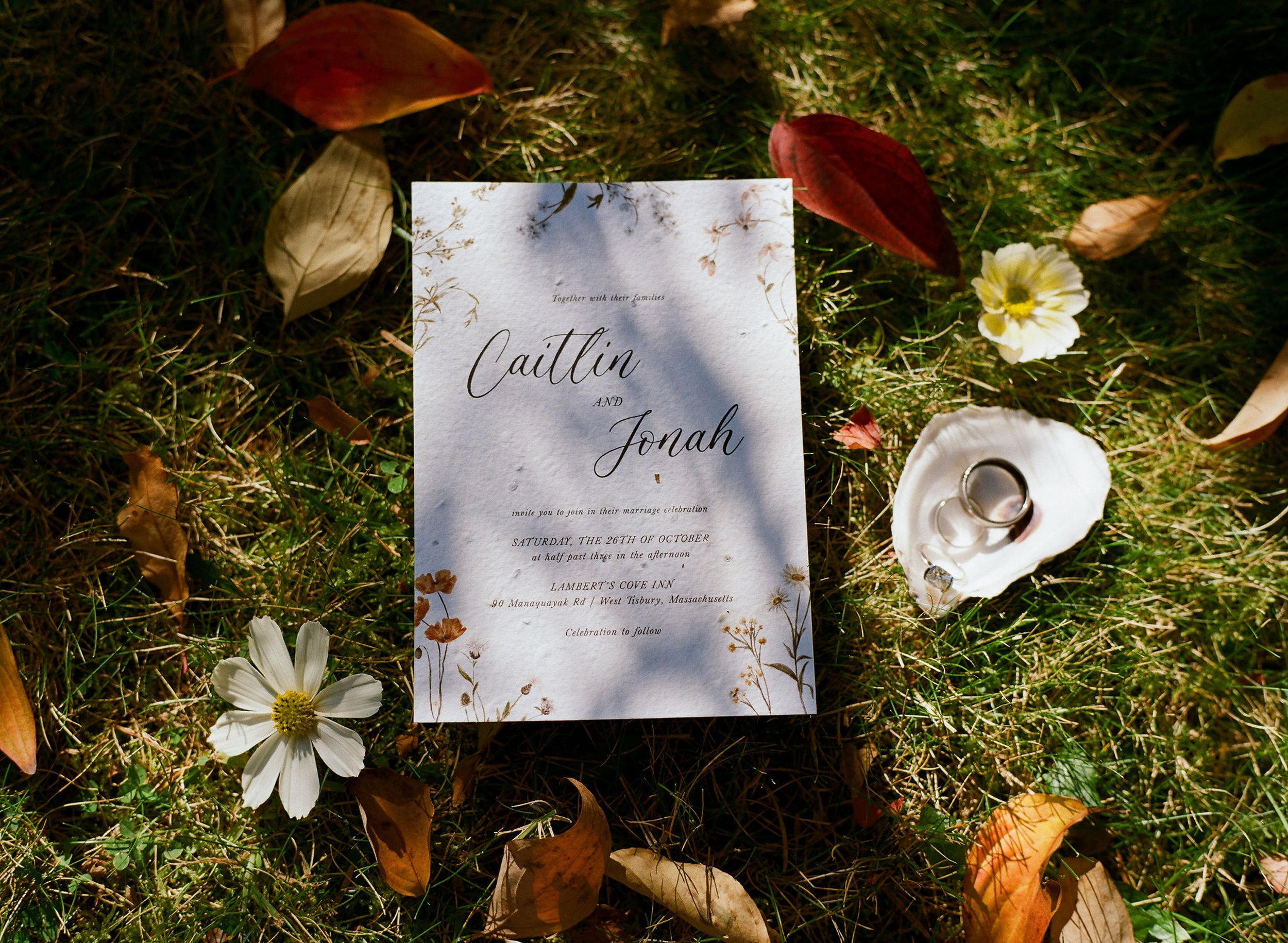 Wedding invitation card on grass surrounded by fallen autumn leaves and white flowers during a wedding at Lambert's Cove Inn on Martha's Vineyard.